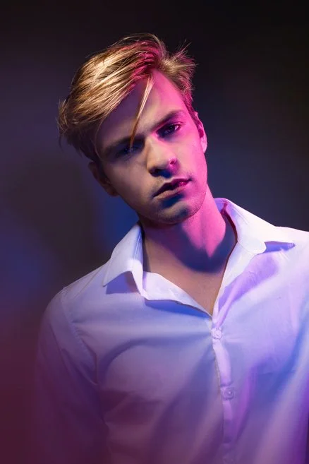 Young man with blond hair wearing a white shirt, illuminated with purple and pink lighting, against a dark background.