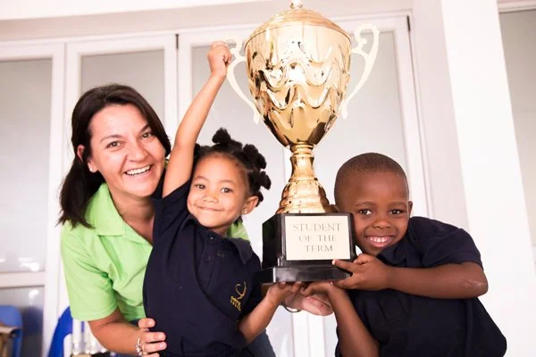 A woman and two children holding a large trophy together in a room with white walls and shelves.