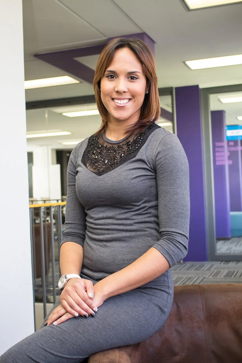 A woman with shoulder-length brown hair, wearing a gray dress with black embellishments on the neckline, sitting on a brown leather chair in an indoor setting with purple walls and ceiling lights.