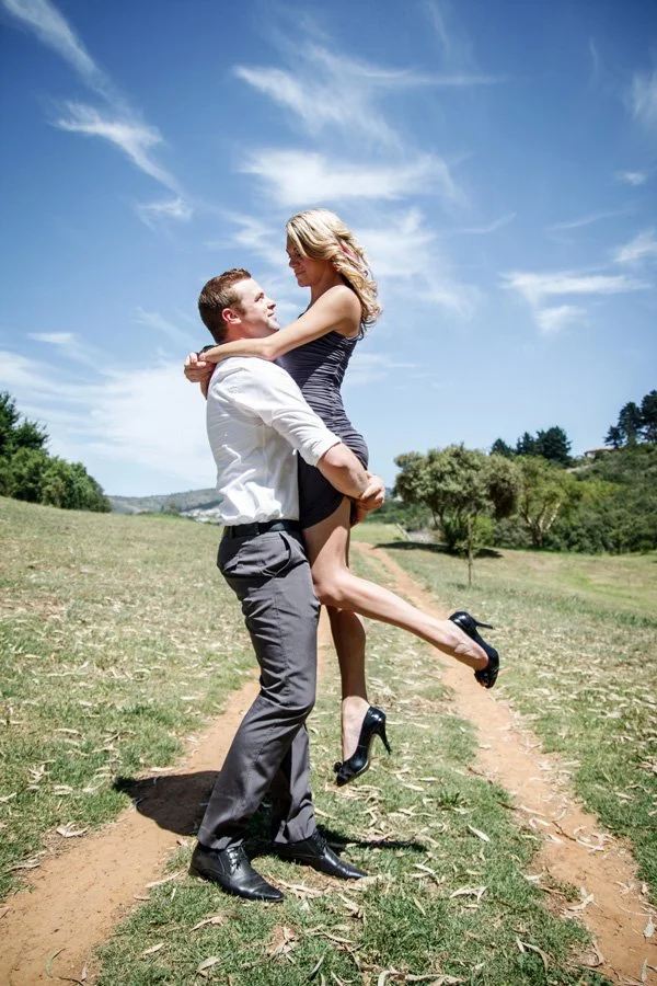 A man lifting a woman in a field with a dirt path, blue sky, and some trees in the background.