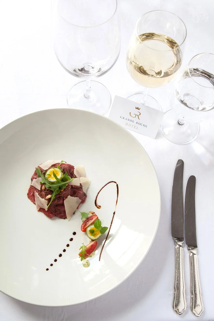 A plated dish of beef carpaccio garnished with arugula, shaved Parmesan cheese, cherry tomatoes, and microgreens, served with a drizzle of balsamic reduction and a green herb sauce. Three glasses of white wine are on the table, along with a hotel nam