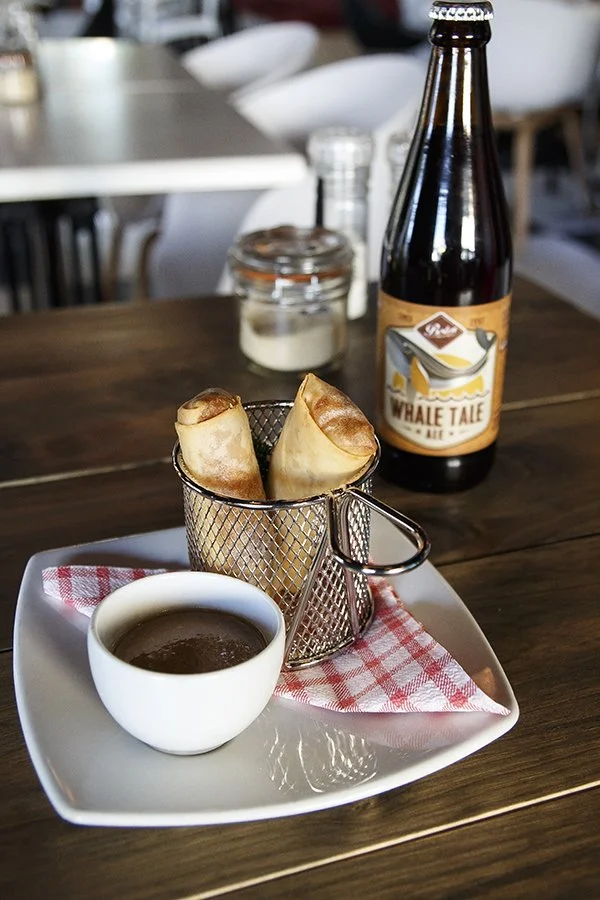 Two egg rolls in a small metal basket, a cup of dipping sauce, a bottle of beer labeled 'Whale Tale Ale', and salt and pepper shakers on a wooden table with a checkered napkin underneath the sauce.