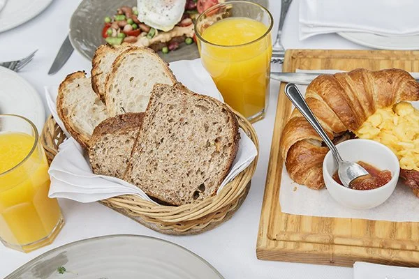 Toast bread, orange juice, scrambled eggs, croissants with jam, and a breakfast salad on a white table.