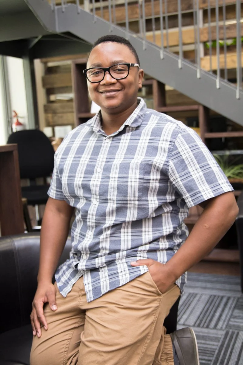 A young man with glasses wearing a blue and white plaid shirt and khaki pants, smiling and posing indoors in front of wooden stairs and furniture.
