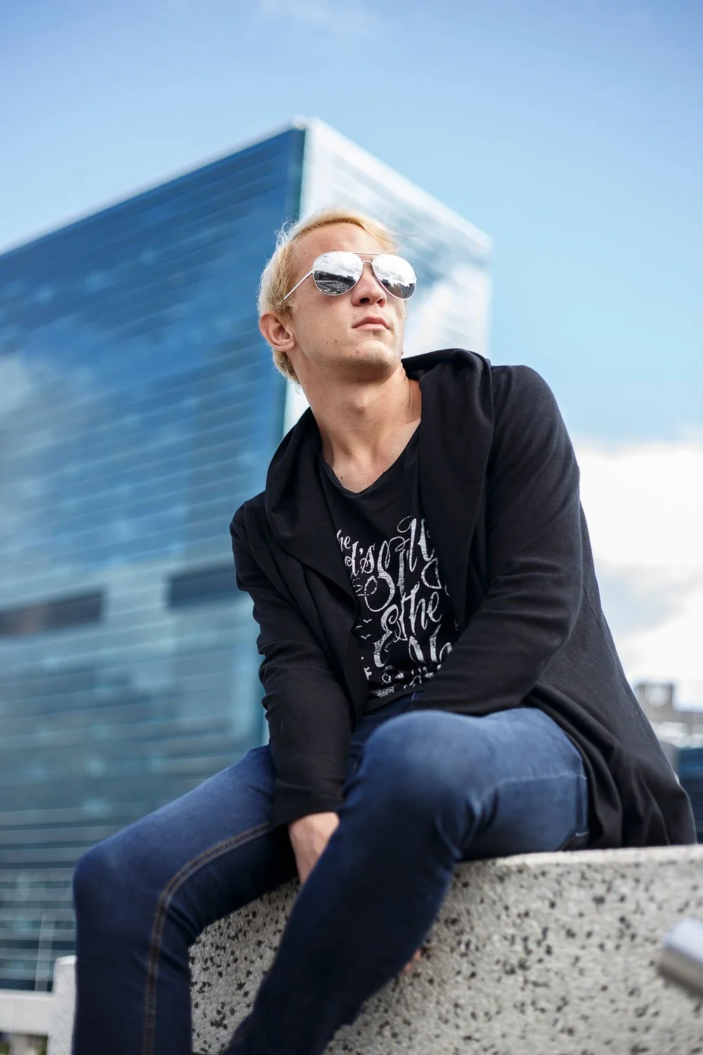 Young man with blonde hair wearing sunglasses, a black hoodie, and a black graphic t-shirt, sitting on a concrete ledge near a modern glass building on a partly cloudy day.