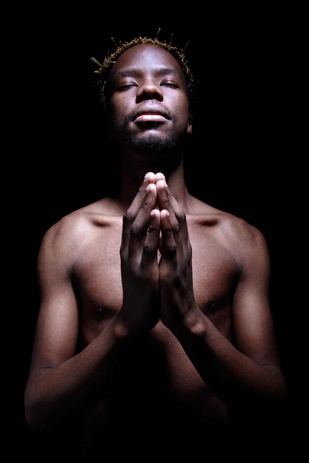 A shirtless man with dark skin stands in darkness with a serious expression, hands pressed together in prayer, wearing a golden crown or headpiece.