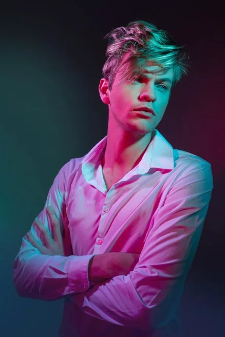 Young man with tousled hair wearing a white button-up shirt, standing against a dark background with colorful pink and teal lighting.