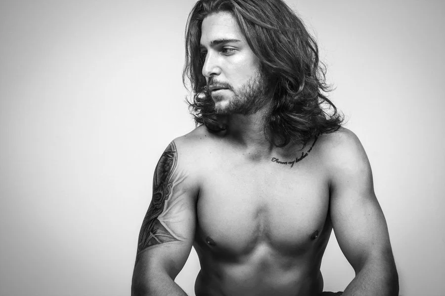 A shirtless man with long wavy hair and a beard, looking to his left, with tattoos on his left shoulder and right chest, in a black and white portrait.