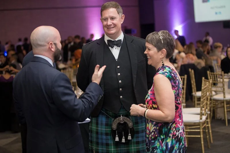 Three people engaged in conversation at a formal event; a man in a tuxedo with a plaid kilt, and a woman in a colorful dress, in a room with many seated guests and purple ambient lighting.