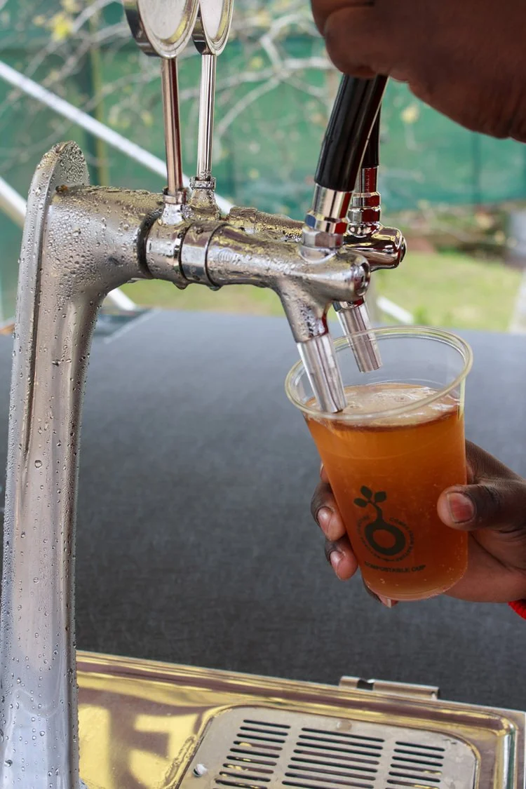 A person fills a clear plastic cup with apple cider from a fountain with a built-in tap. The cup has a black print of a tap and text underneath. Water droplets are on the metal fountain.