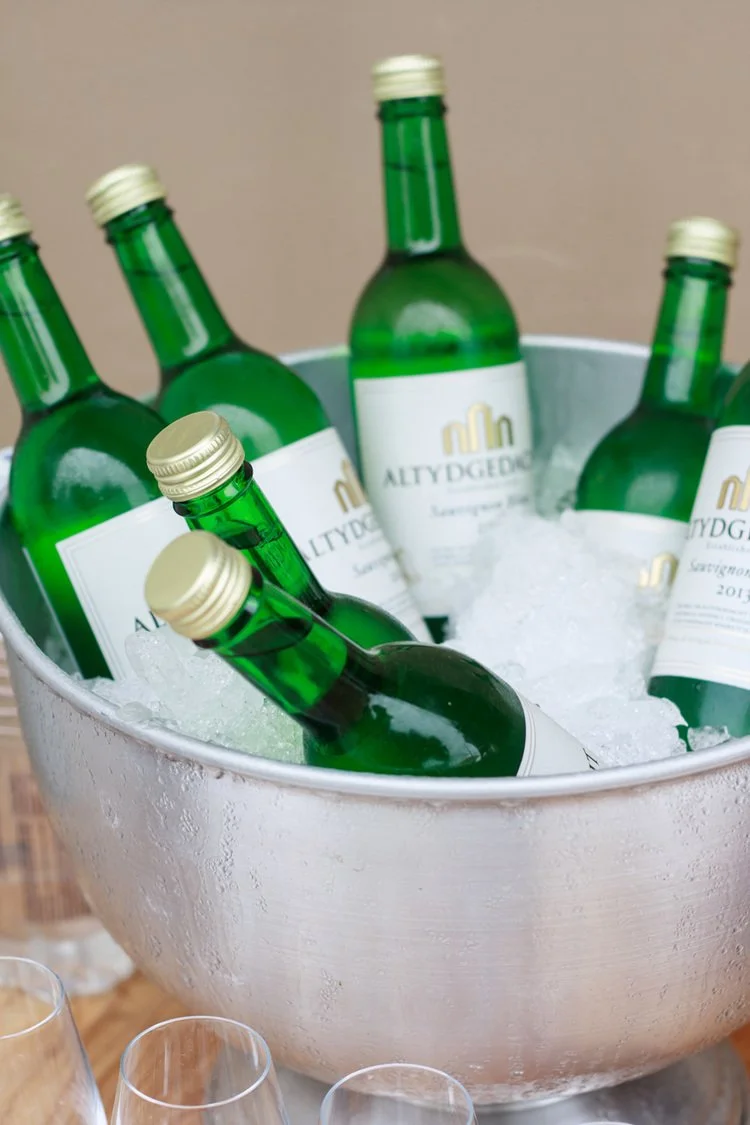 Green glass bottles of ALTYDODGE wine chilling in a large metal ice bucket filled with ice.