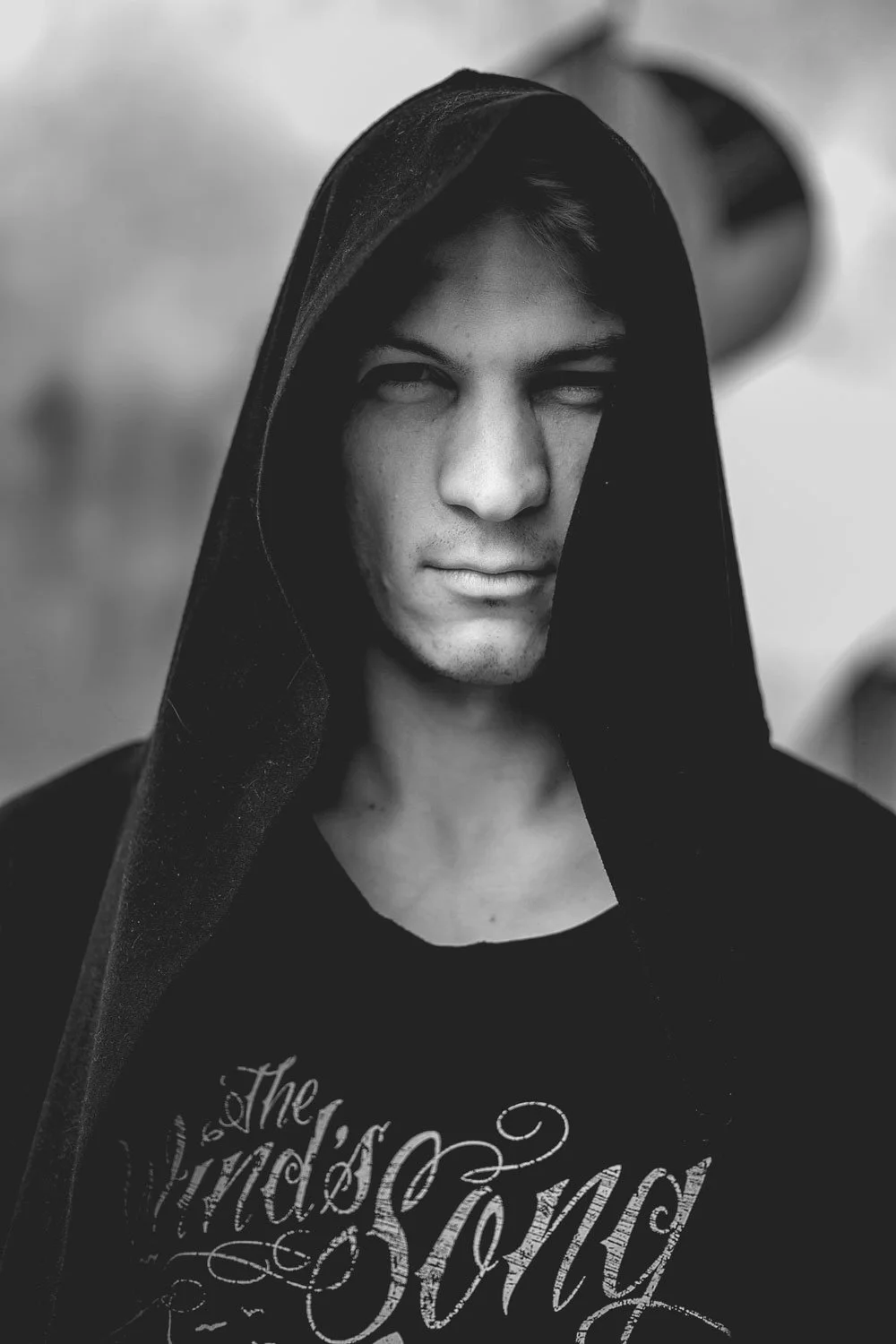 Black and white portrait of a young man with a serious expression, wearing a black hooded garment and a T-shirt with artistic text on it.
