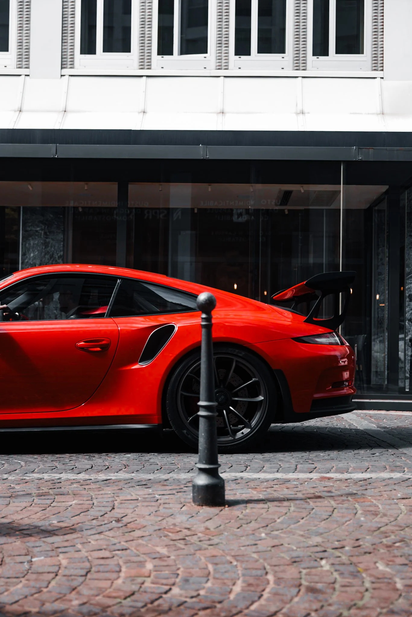 Red sports car parked on cobblestone street in front of modern building with large windows and white facade.