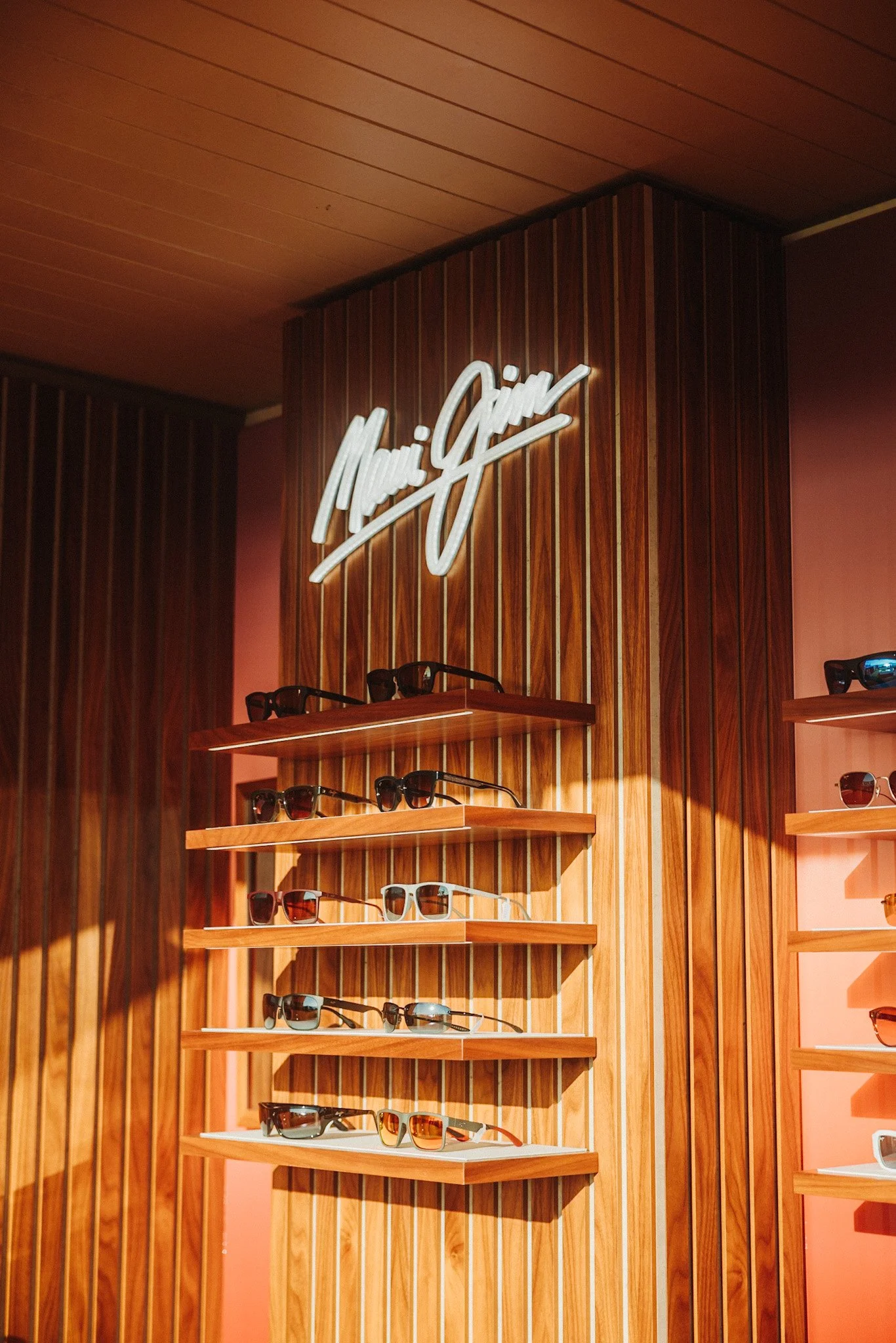 Display of various sunglasses arranged on wooden shelves inside a store with a neon sign reading 'Maui Jim' on a wooden-paneled wall.