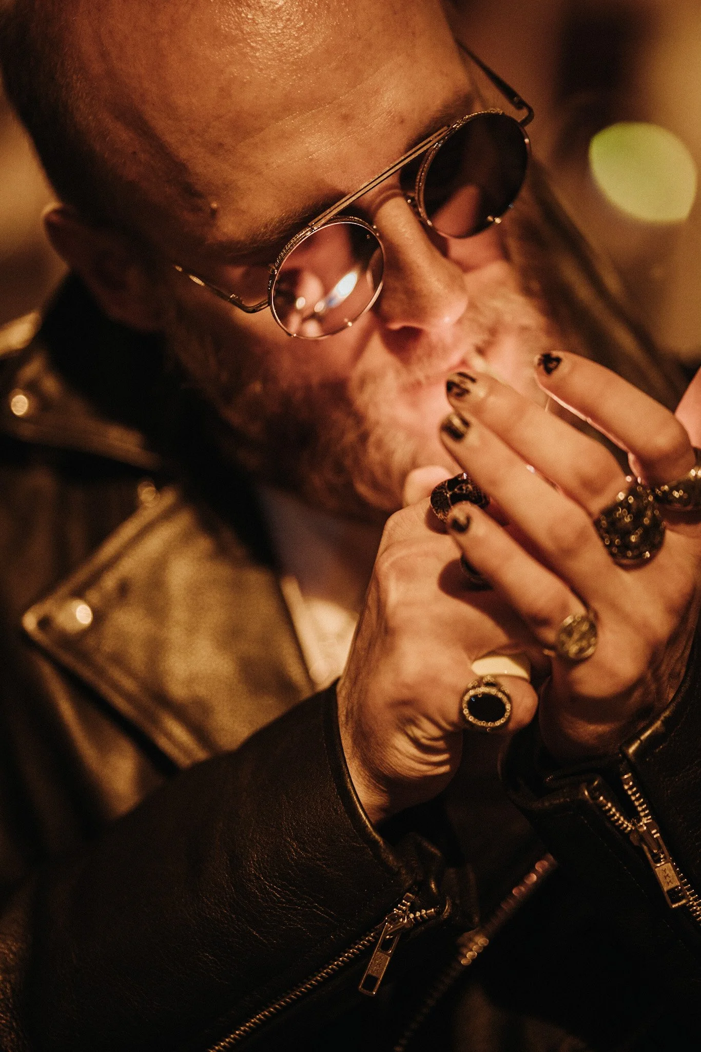 Close-up of a person wearing round sunglasses, with rings and black nail polish, in a dimly lit environment.