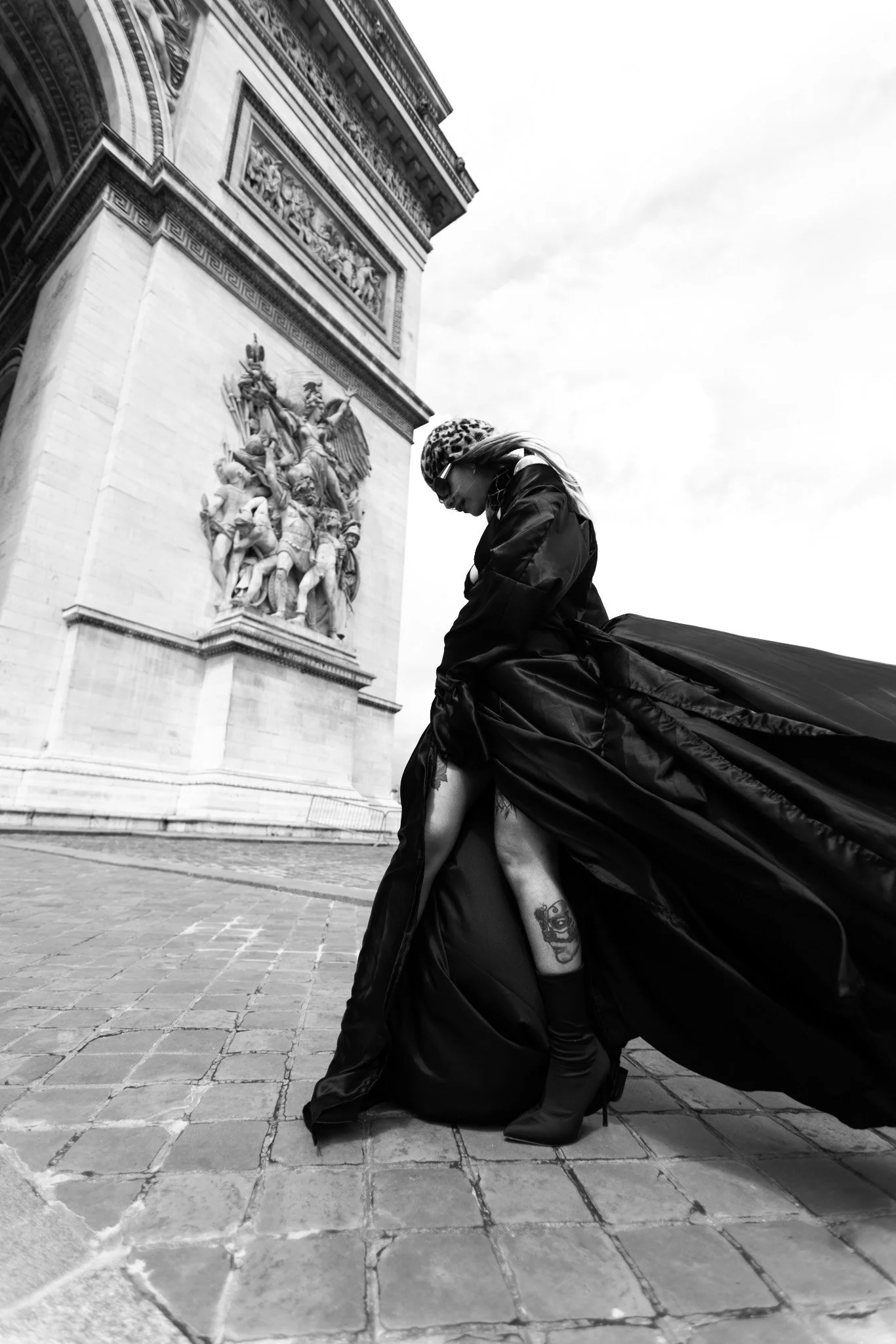 A woman in high heels and a long flowing black dress with a tattoo on her thigh, standing on cobblestone street in front of a monument with detailed sculptures, looking down with her head slightly bowed, wearing sunglasses and a patterned headscarf, 