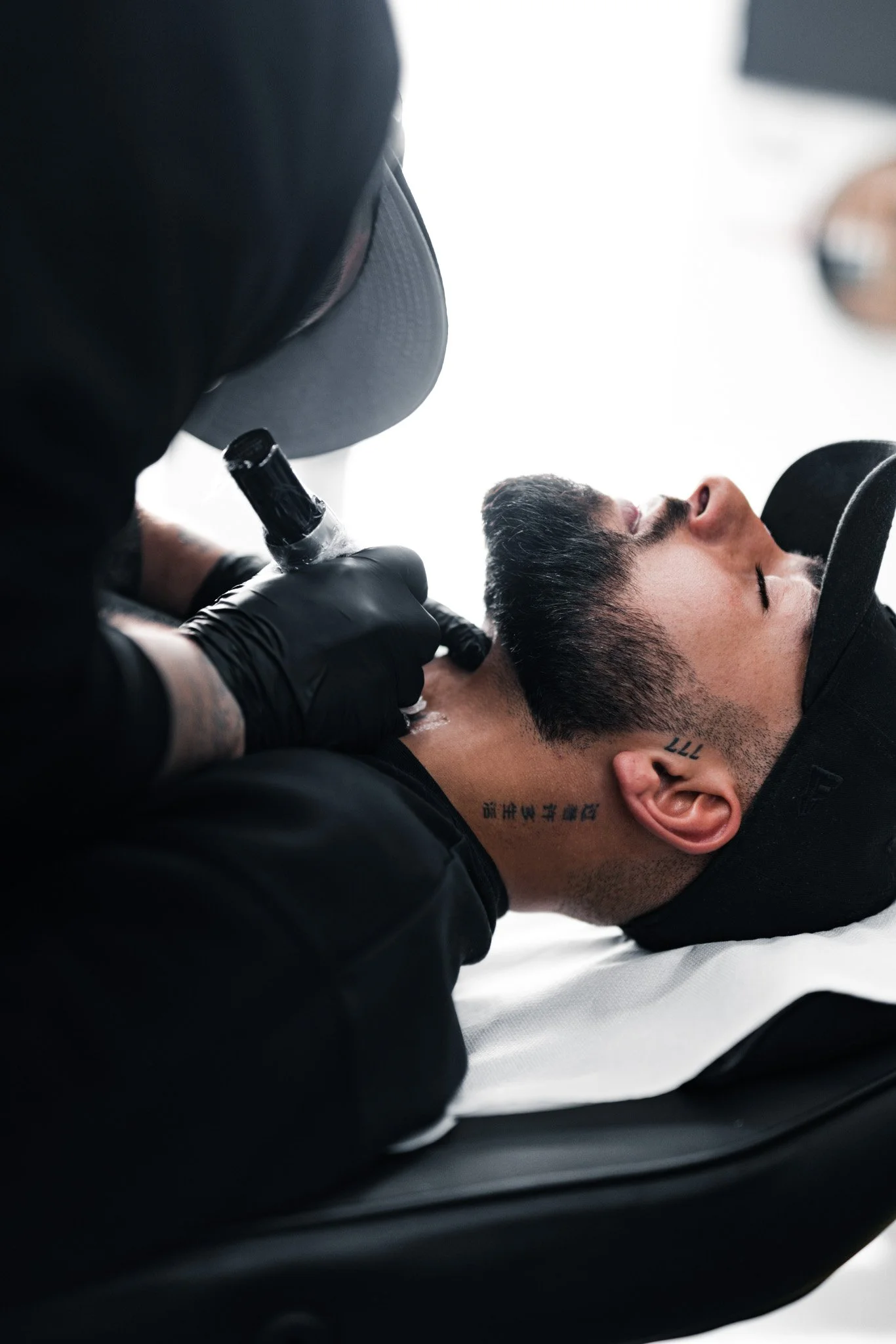 Tattooed man lying down with eyes closed, receiving a tattoo on his neck from a tattoo artist wearing black gloves in a well-lit room.