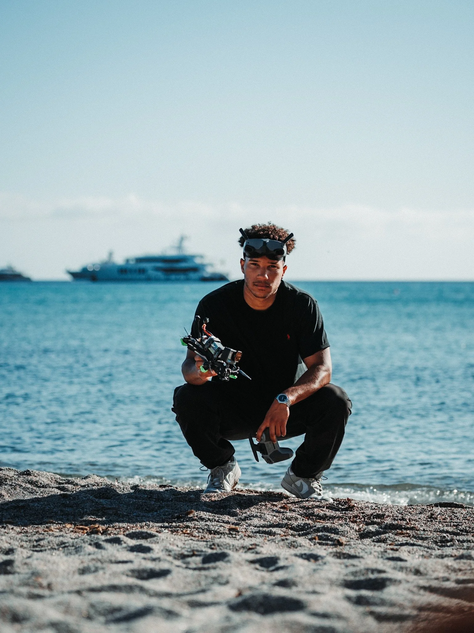 Young man crouching on a sandy beach near the water, wearing a black t-shirt, gray sneakers, and holding a drone controller with goggles on his head. A yacht is visible in the background on the water.