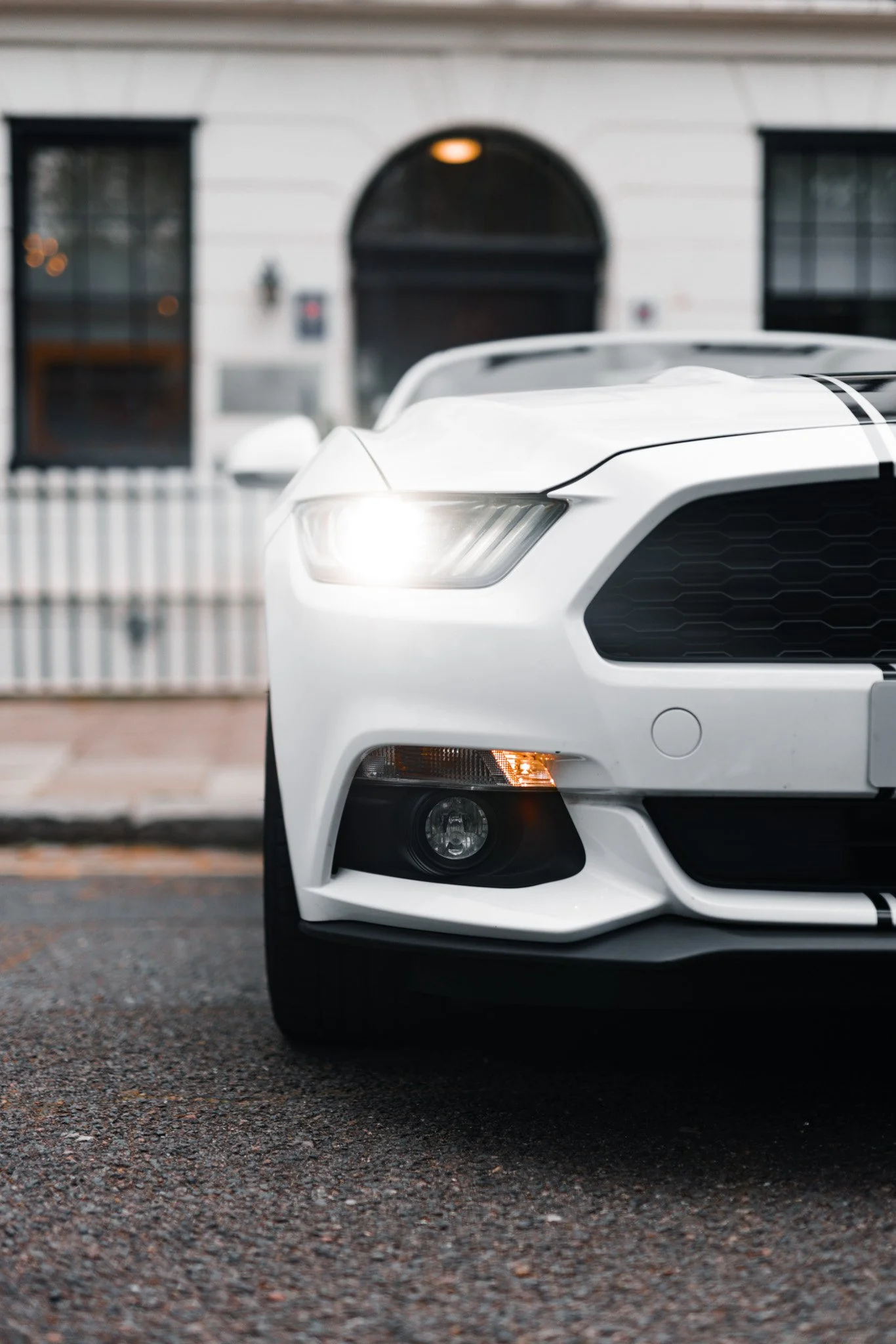 Close-up of the front white sports car with a black grille, headlight, and fog light, parked on a paved surface in front of a white house with black shutters and a black arched door.