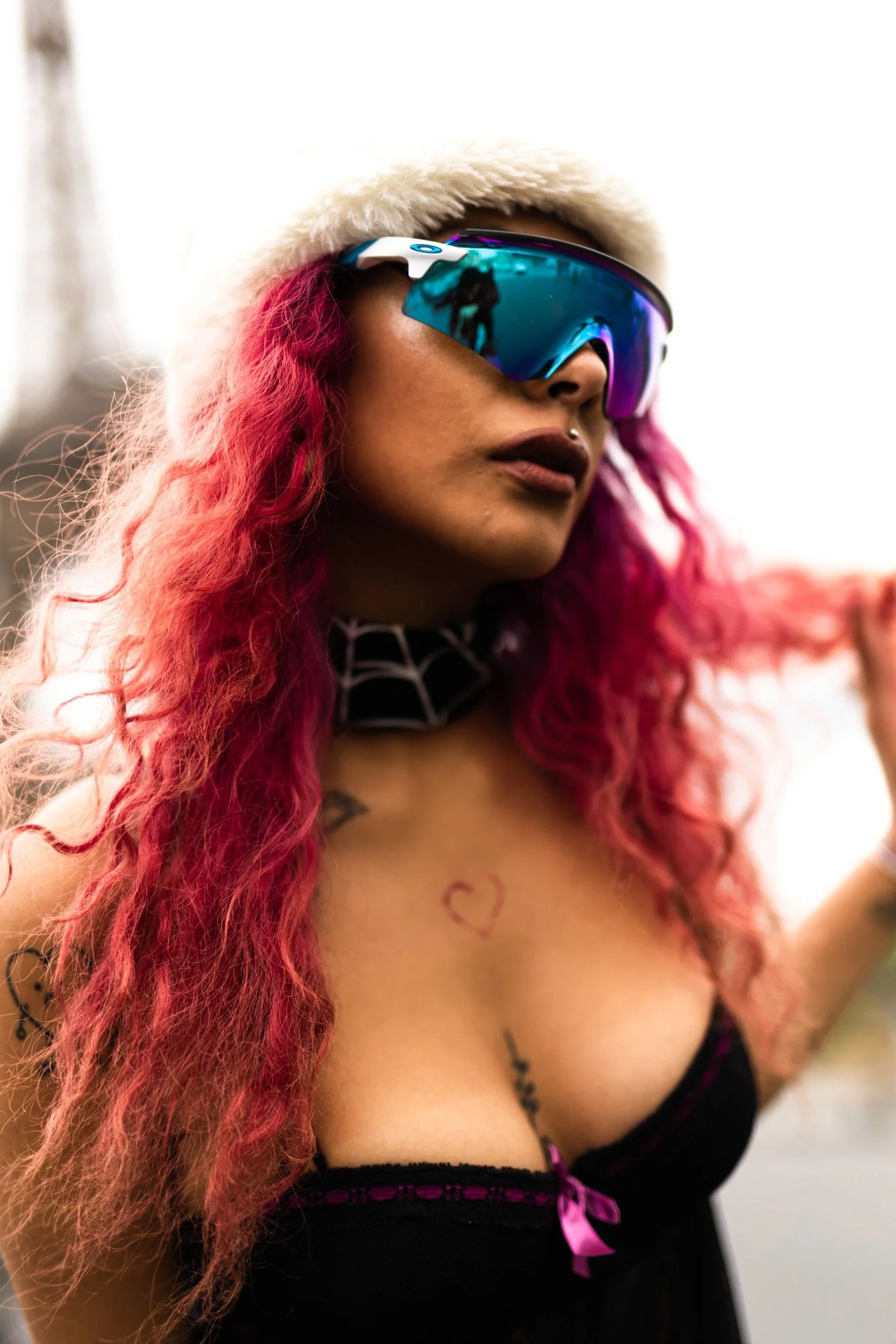 A woman with long, curly pink hair wearing a white furry hat and reflective, sports-style sunglasses. She has a piercing above her upper lip and tattoos on her shoulder and chest. She is wearing a black garment with pink ribbons.