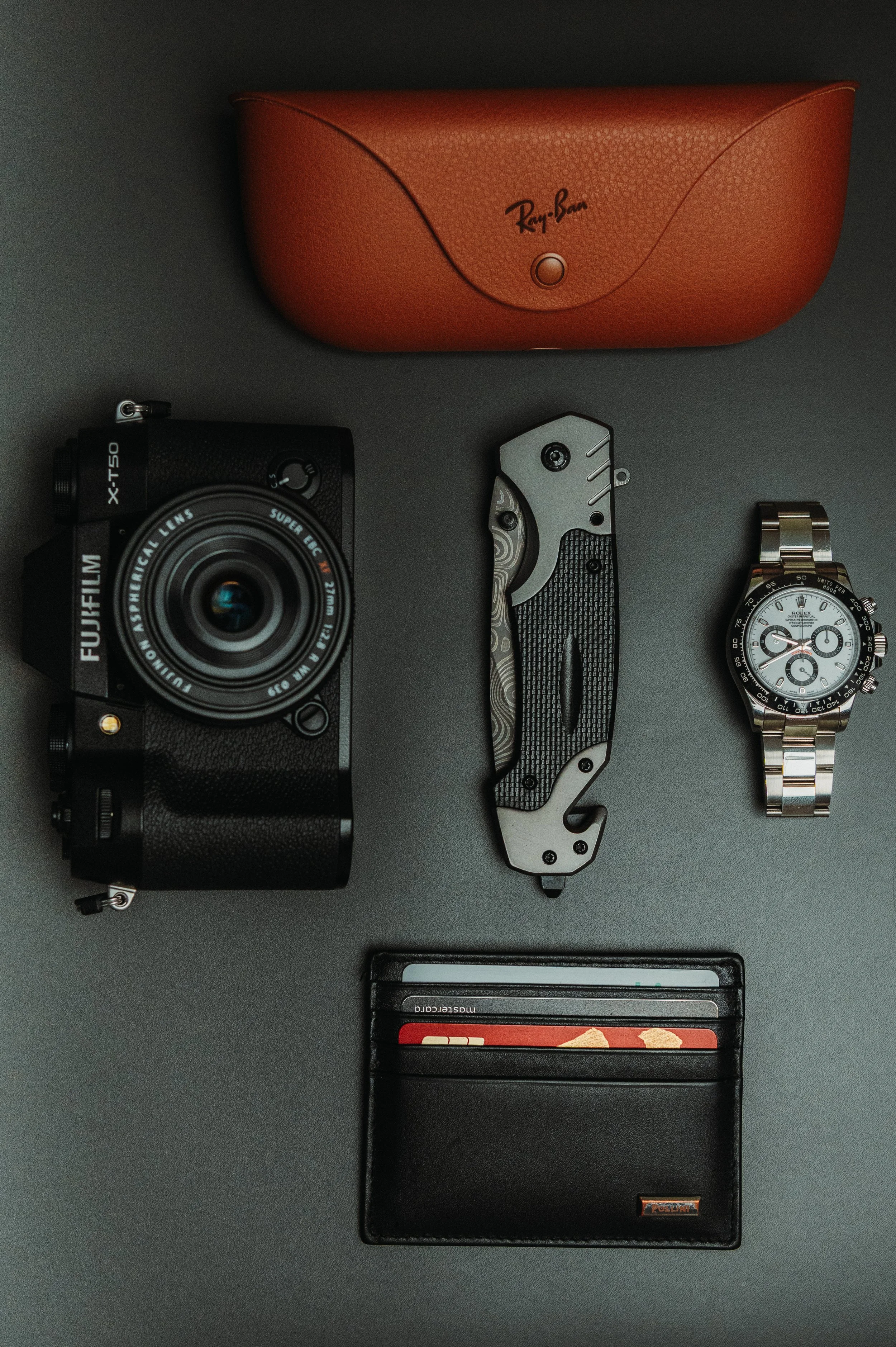 A collection of accessories and electronics on a gray surface, including a vintage film camera, a folding pocket knife, a wristwatch, a leather case, and a small container with red and black pens or markers.