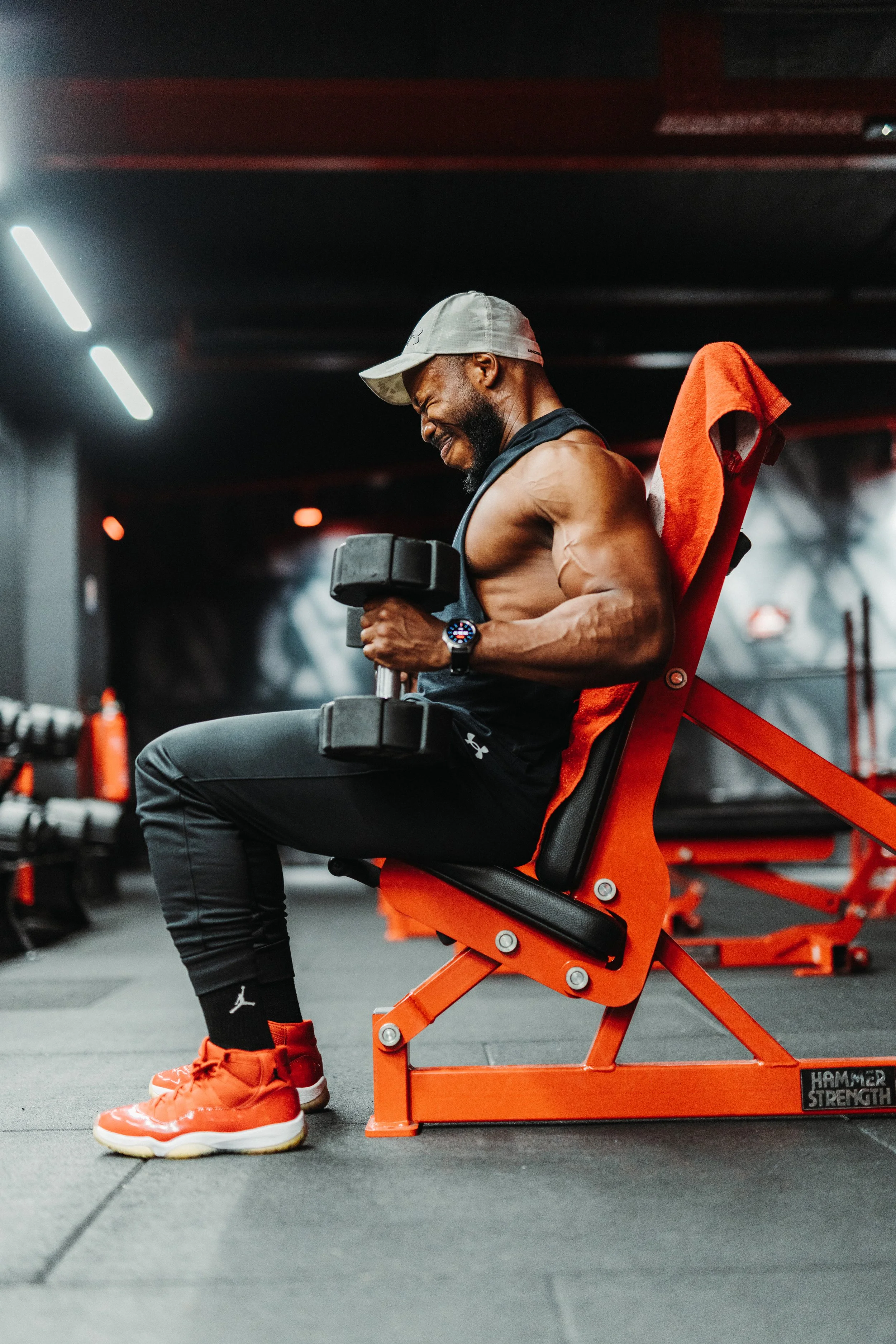 Man sitting on an orange workout bench in a gym, lifting a dumbbell with a smile, wearing a gray cap, black sleeveless shirt, black athletic pants, bright orange shoes, and a watch, with gym equipment blurred in the background.