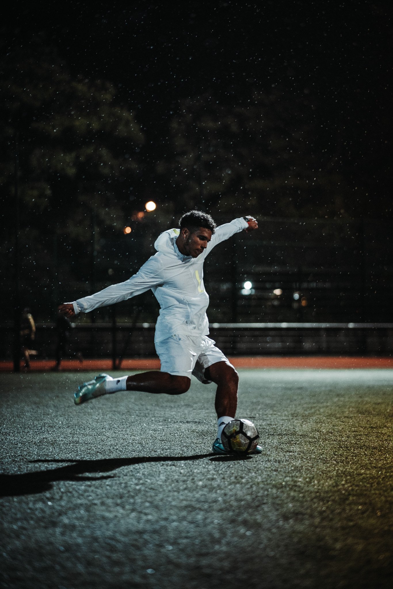 A man in a white sports outfit is playing soccer on a wet outdoor court at night, with raindrops falling around him, and street lights illuminating the scene.