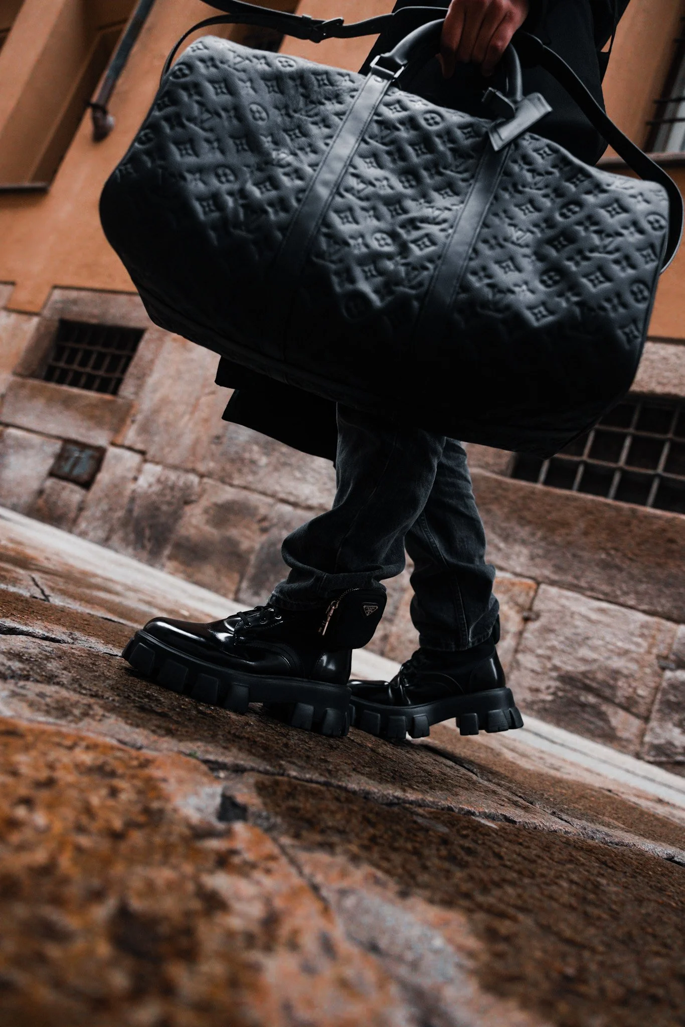 Person walking on cobblestone street carrying a black designer duffel bag with embossed logo pattern, wearing black platform shoes, black jeans, and a dark jacket.