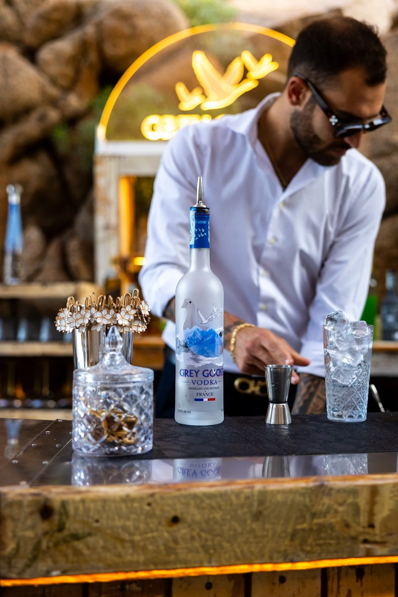 A bartender wearing sunglasses and a white shirt preparing drinks at a bar with a wooden countertop. There are various bottles, glassware, and a neon sign with a dove in the background.