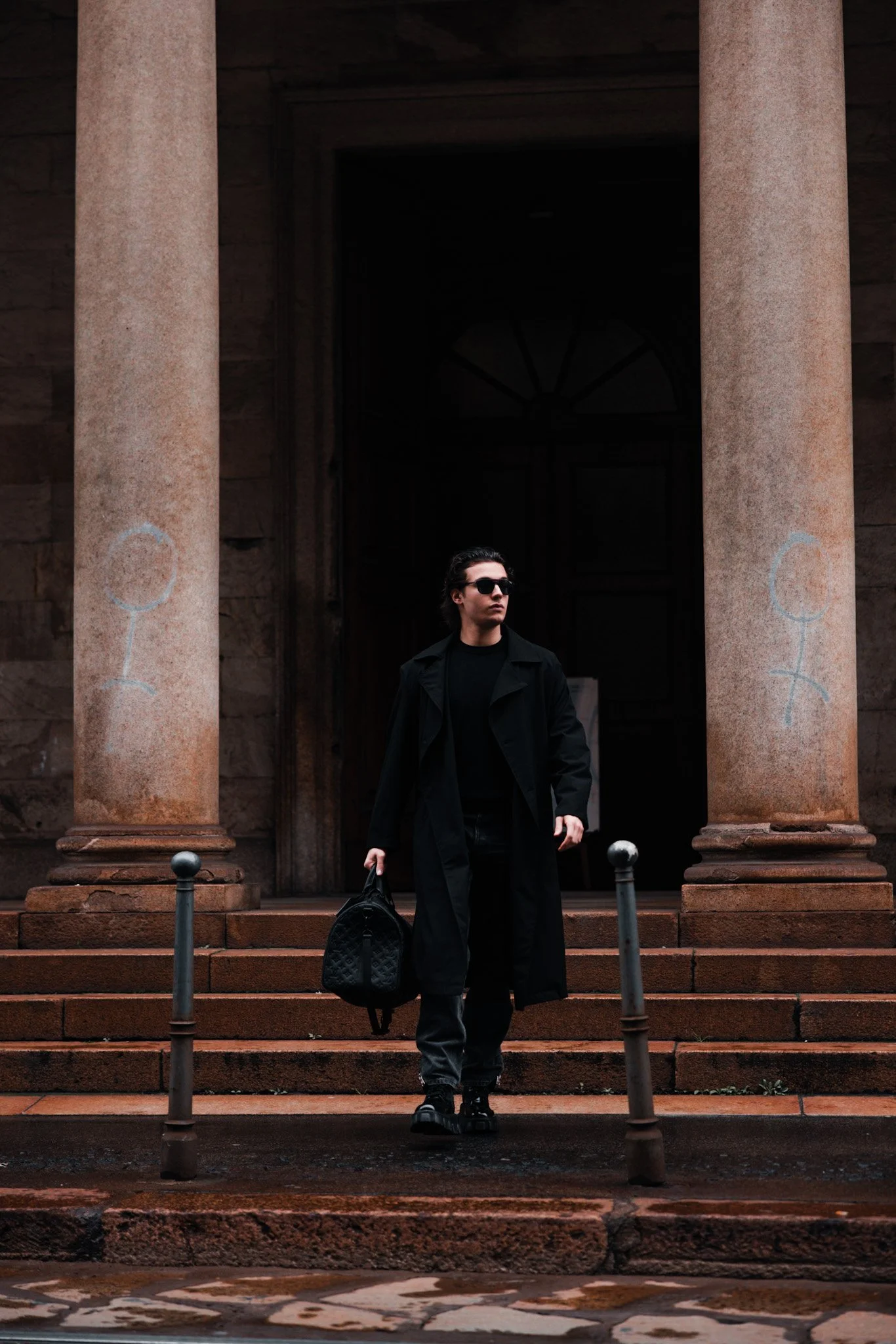 A young man with sunglasses, wearing a black coat and pants, walking down the stairs of a historical building with large stone columns, holding a black bag.