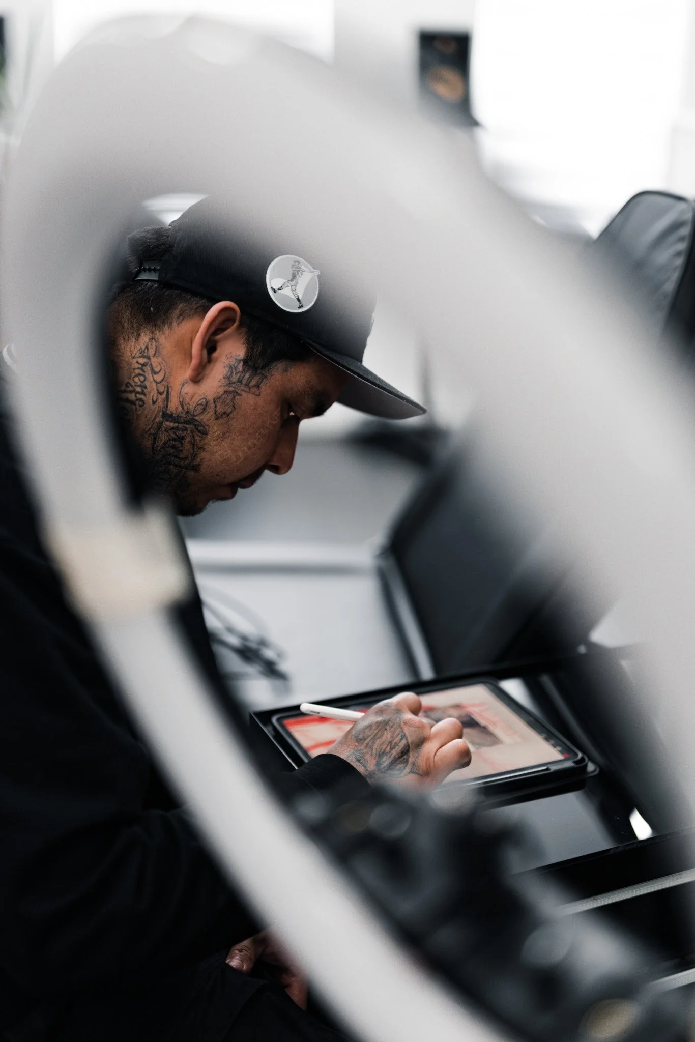 A person with tattoos on their neck and hand, wearing a black cap with a logo, using a stylus on a tablet.
