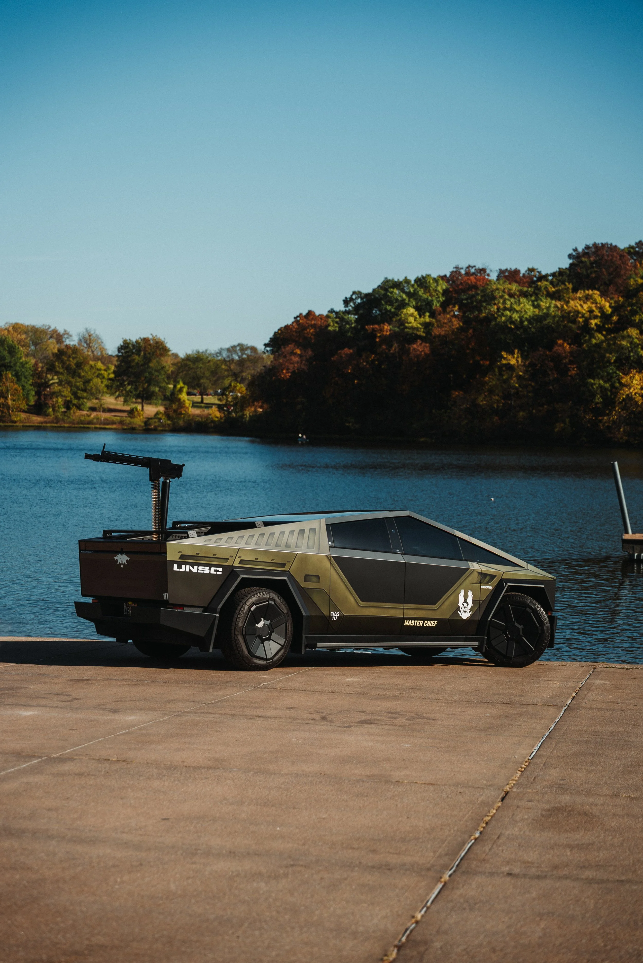 A futuristic military-style vehicle parked on a concrete area beside a body of water, with trees showing fall foliage in the background under a clear blue sky.