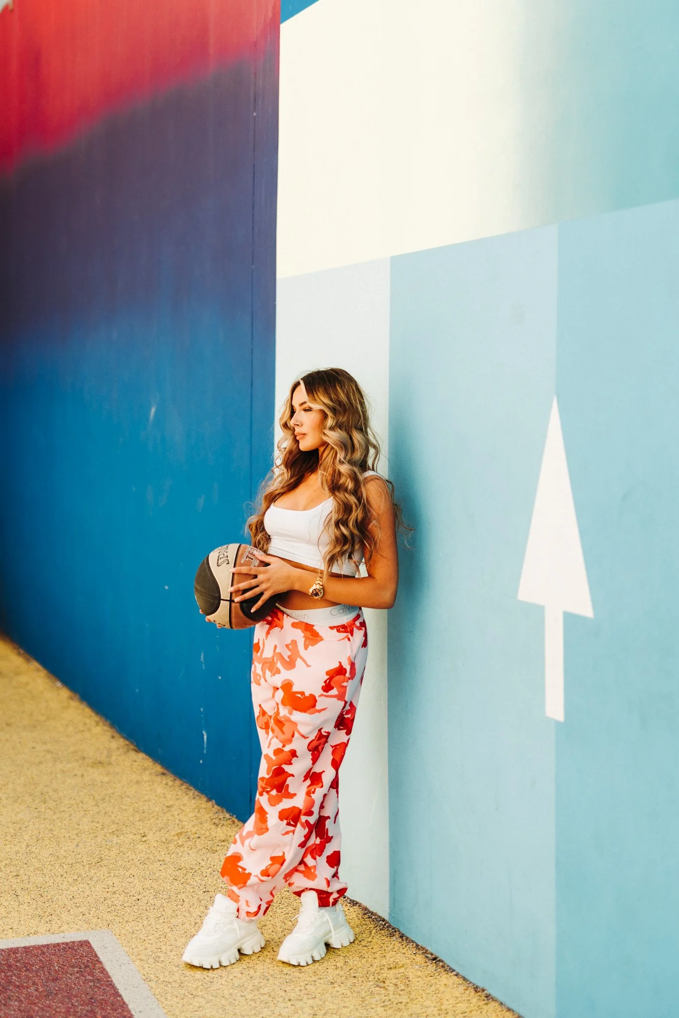 A woman with long wavy hair, wearing a white crop top, floral pants, and chunky white sneakers, is standing against a blue wall with a white geometric arrow sign, holding a basketball in her arms.