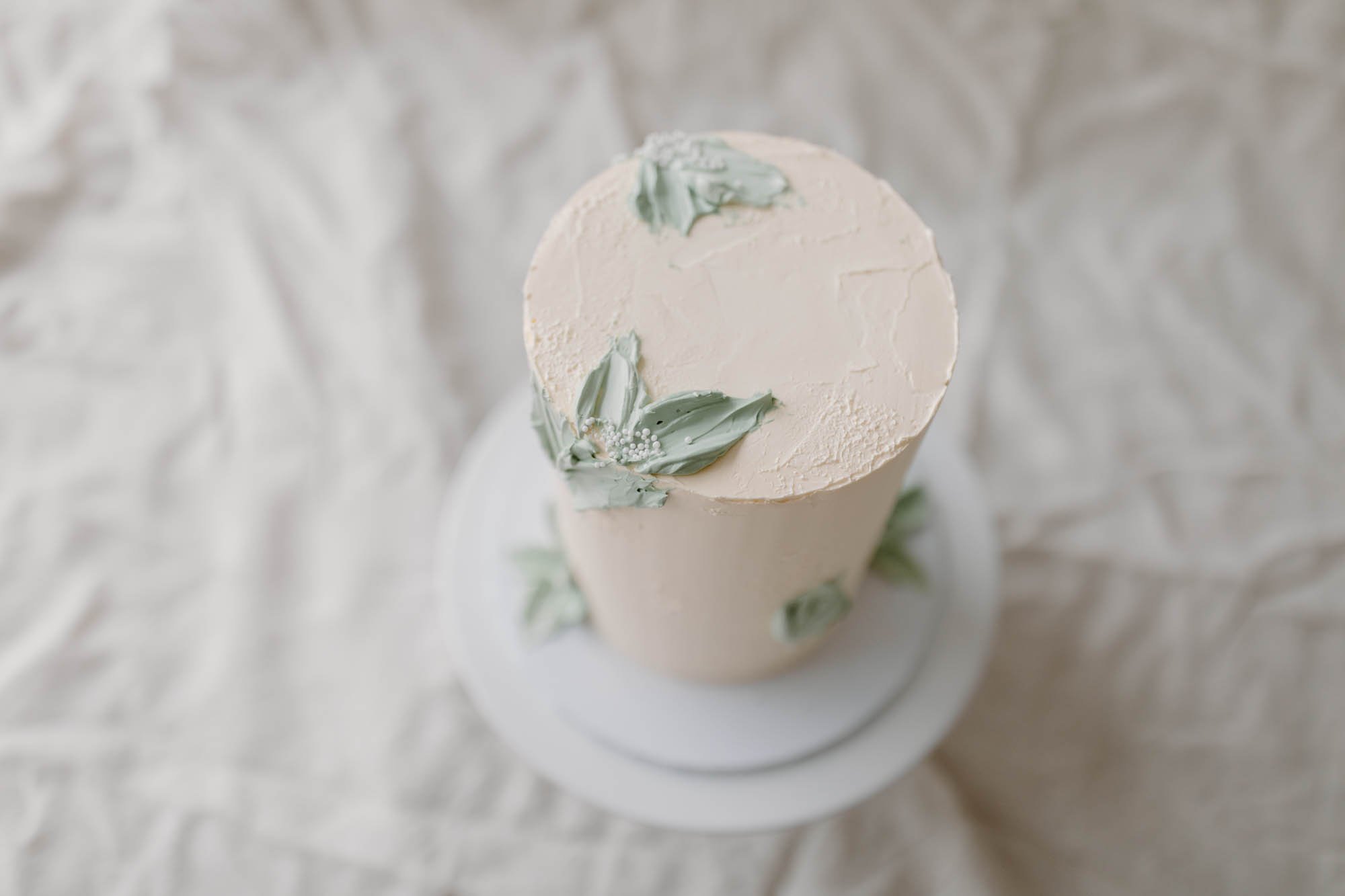 Top view of a tall, cylindrical white cake decorated with green leaves and small white edible beads on a white plate, placed on crinkled white fabric.