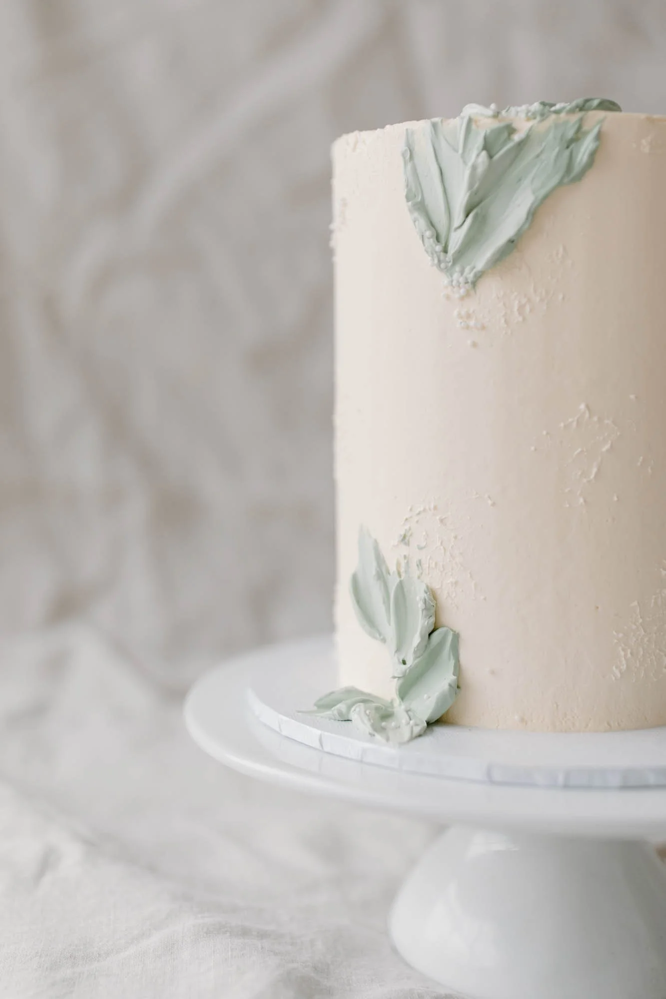 A tall white cake on a white cake stand with light green decorative leaves or frosting applique on the side, set against a textured light grey background.