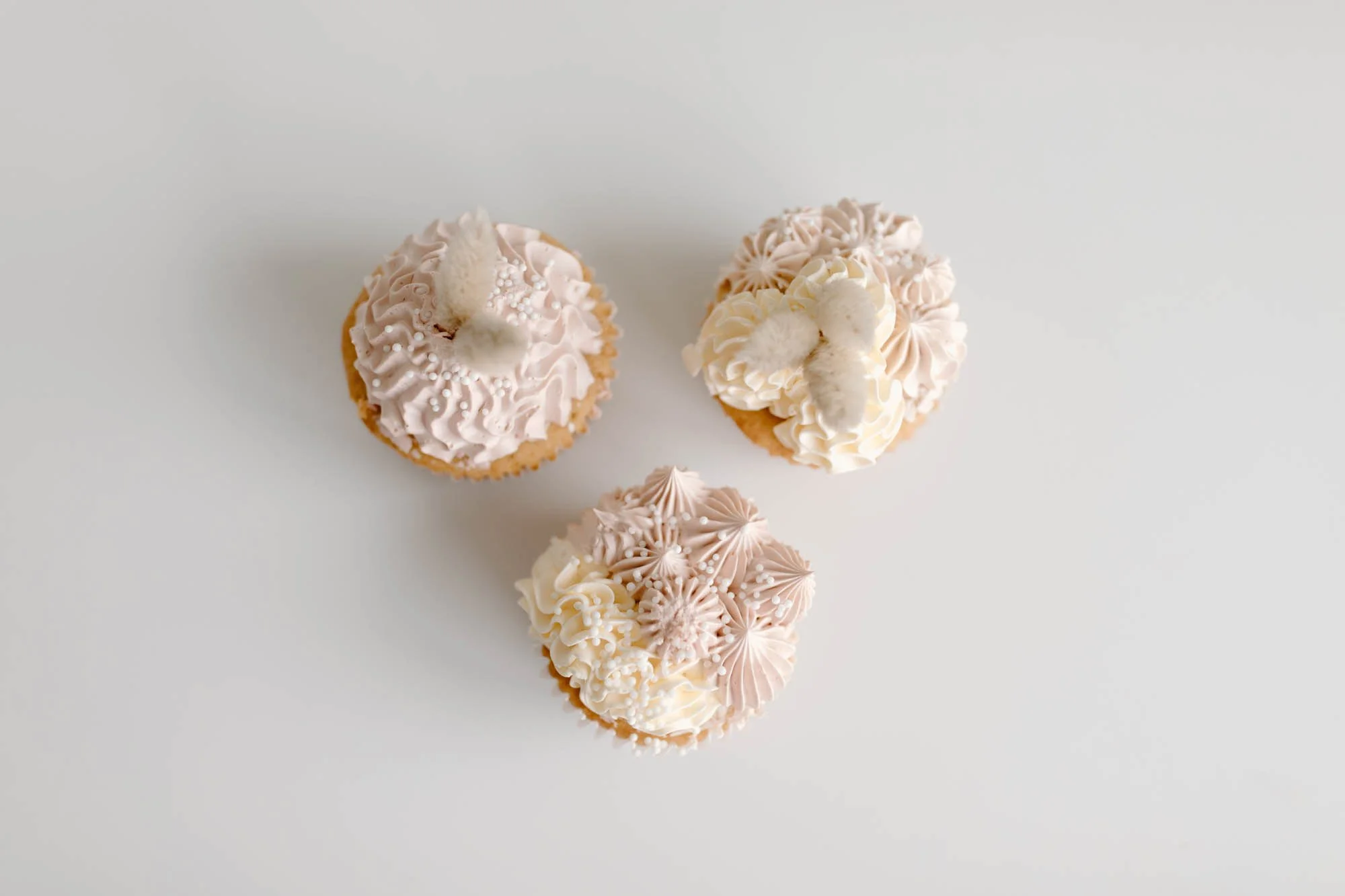 Three cupcakes decorated with pink and white frosting and white sprinkles, arranged on a white surface.