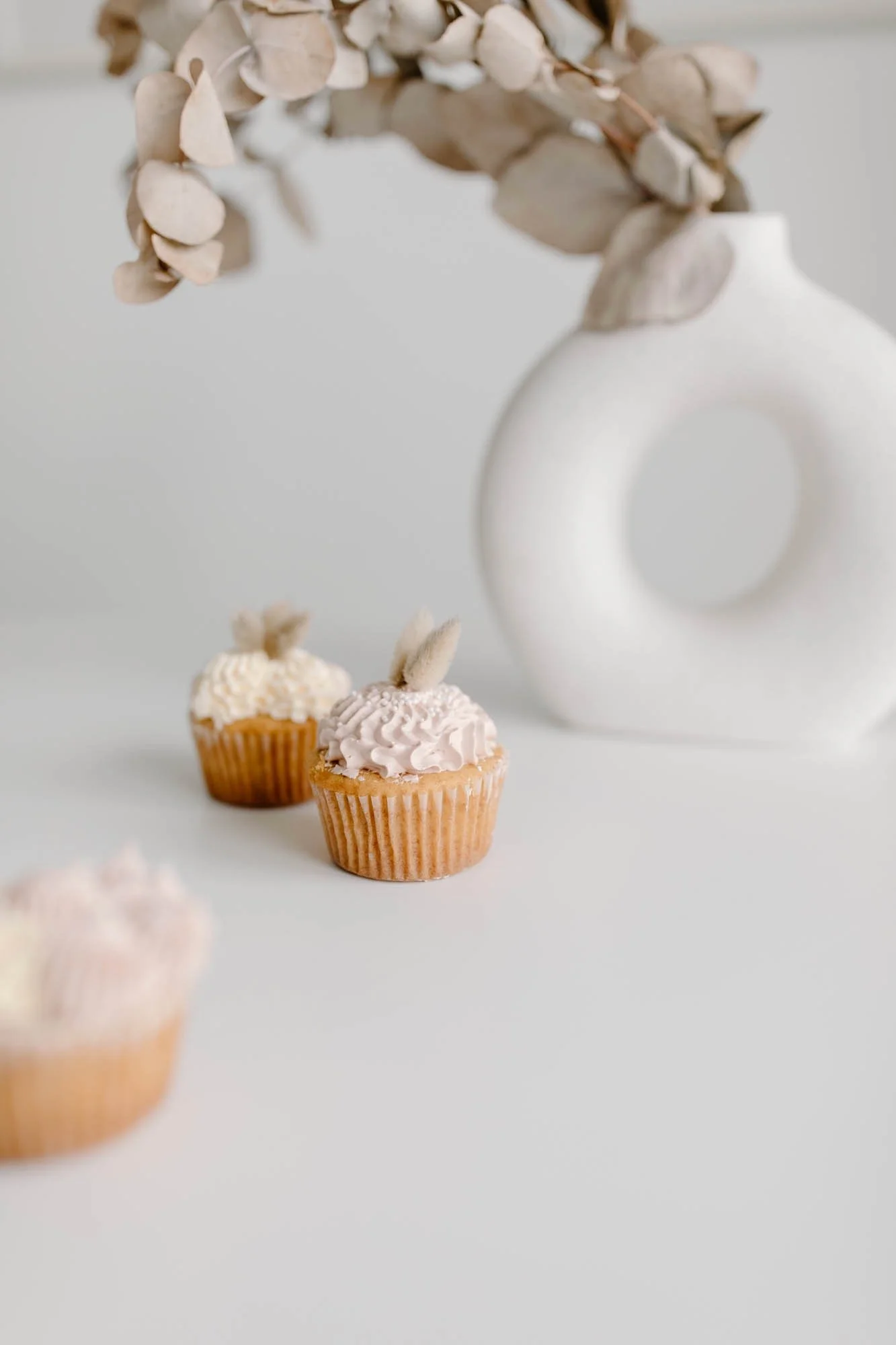 Three cupcakes with pink and white frosting, topped with small plush flowers, placed on a white surface. In the background, there is a white ceramic vase with dried leaves or flowers.