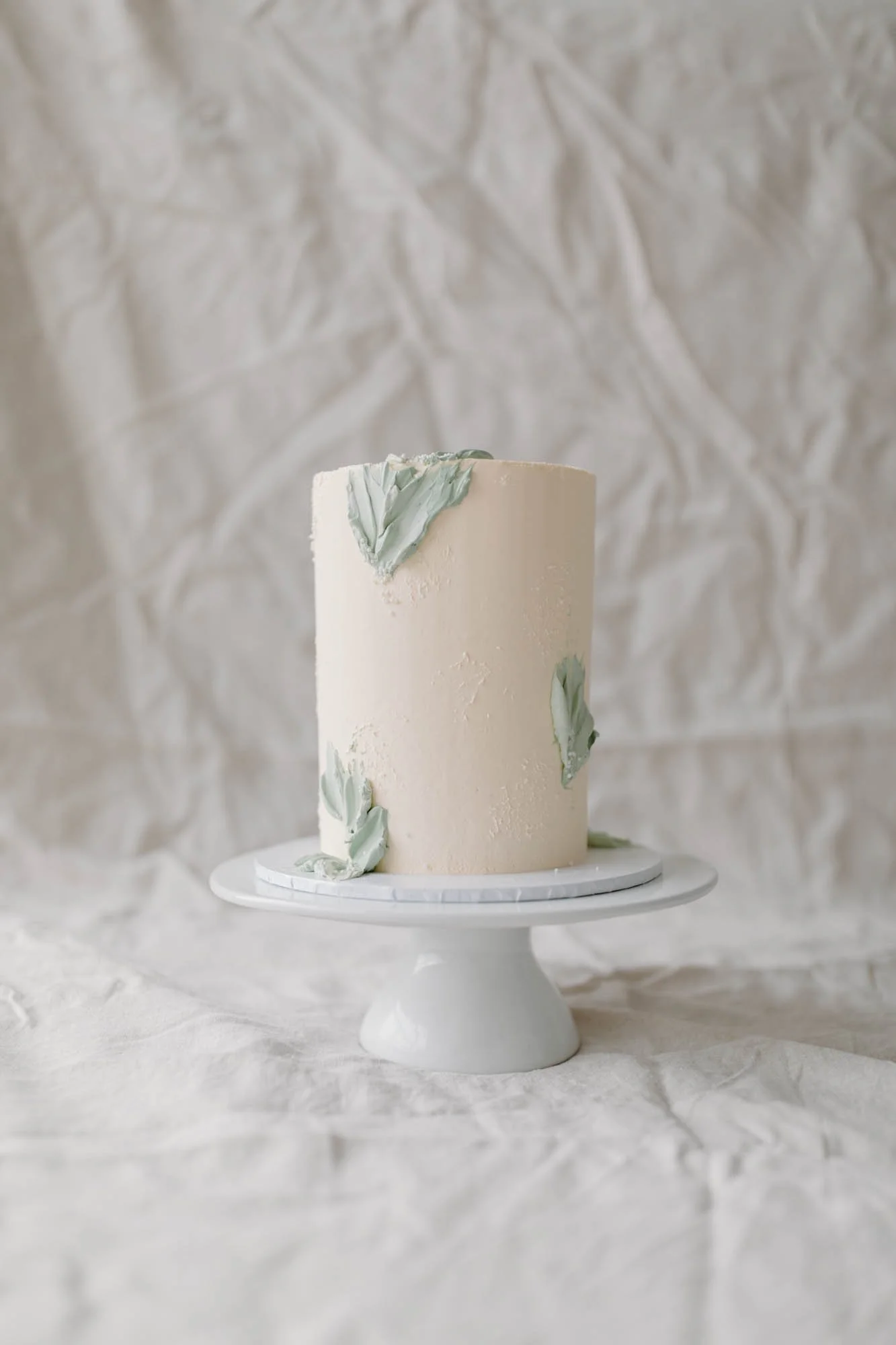 A tall, white, cylindrical cake with light green decorative flowers on a white cake stand in front of a textured, crumpled white background.