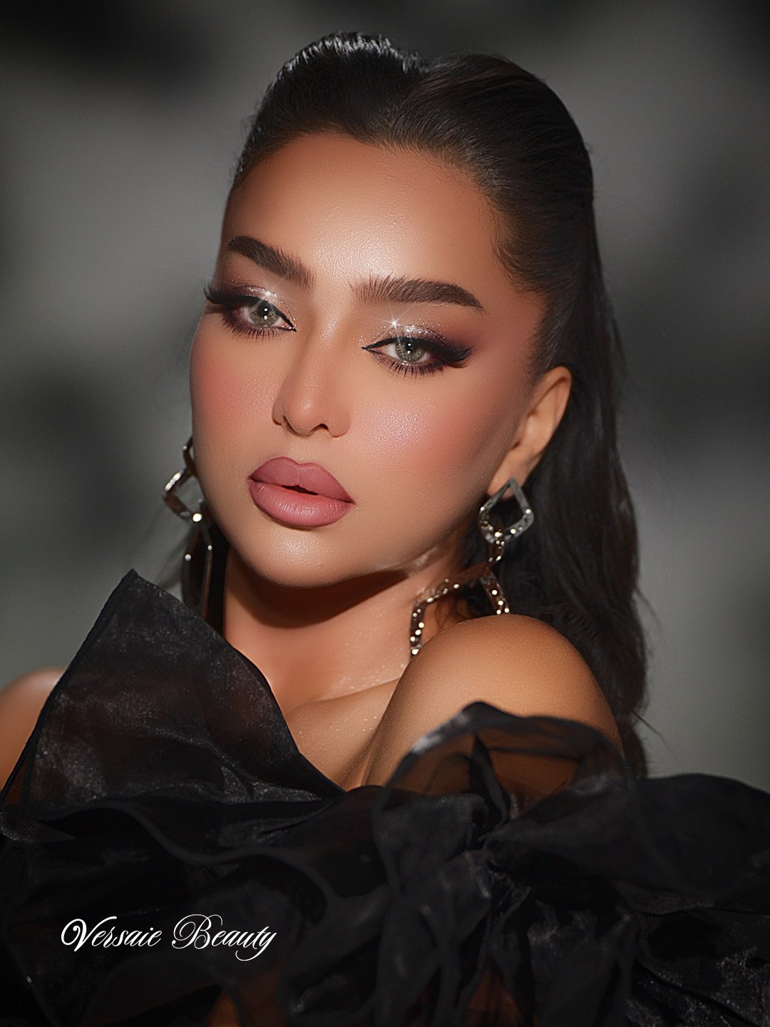 A woman with dark hair styled in waves, wearing glamorous makeup with glitter eyeshadow and nude lipstick, large hoop earrings, and a black off-the-shoulder dress with ruffled sleeves, posing against a gray background.