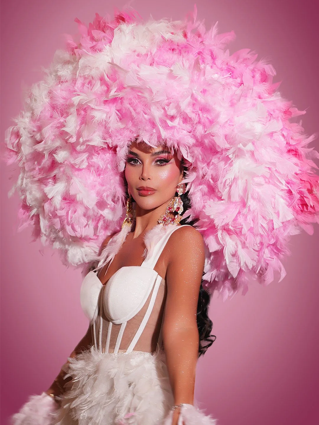 A woman wearing elaborate pink and white feathered headdress, white dress with sheer panels, and large dangling earrings, posed against a pink background.
