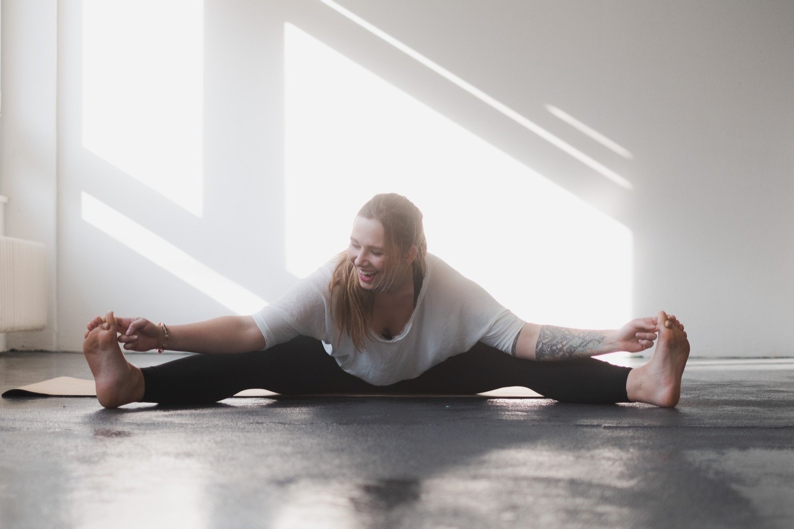 Eine Frau in sportlicher Kleidung macht auf einer Yogamatte eine Dehnübung in einem hellen Raum.