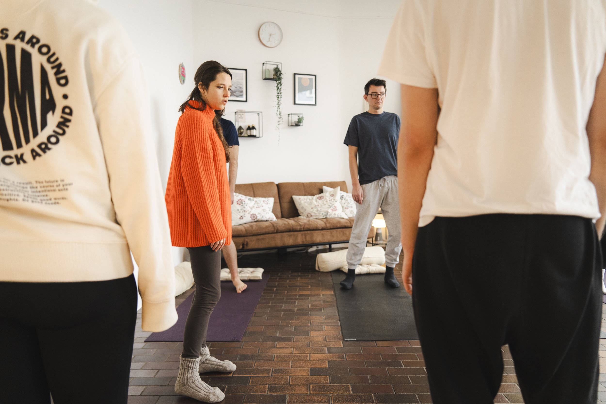Personen in einem Raum auf Yogamatten stehend, im Hintergrund ein Sofa und Wanddekoration.