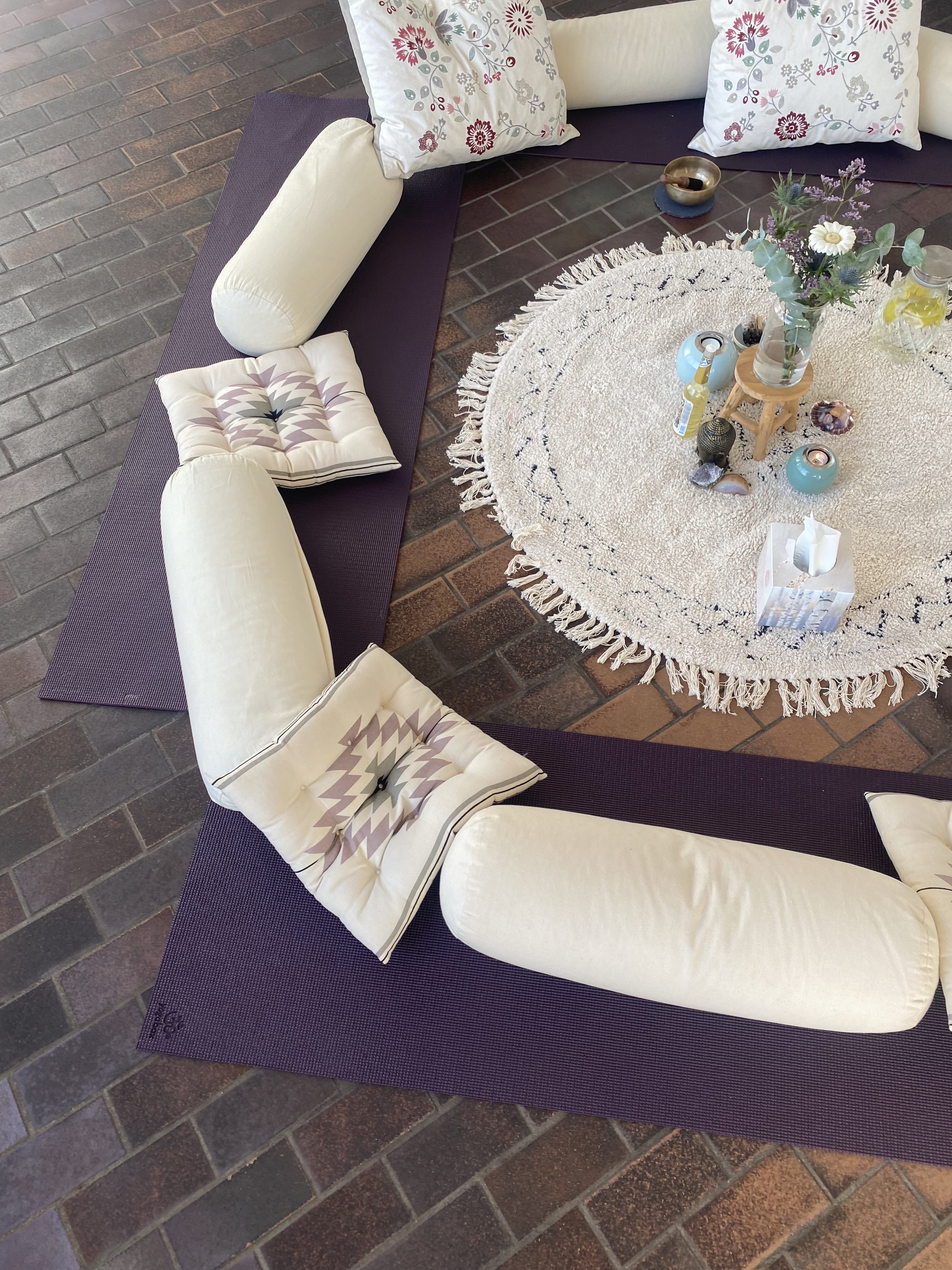 Runde Sitzgelegenheit mit Yogamatten und Kissen auf einem Teppich, dekoriert mit einem Blumenarrangement und Kerzen.