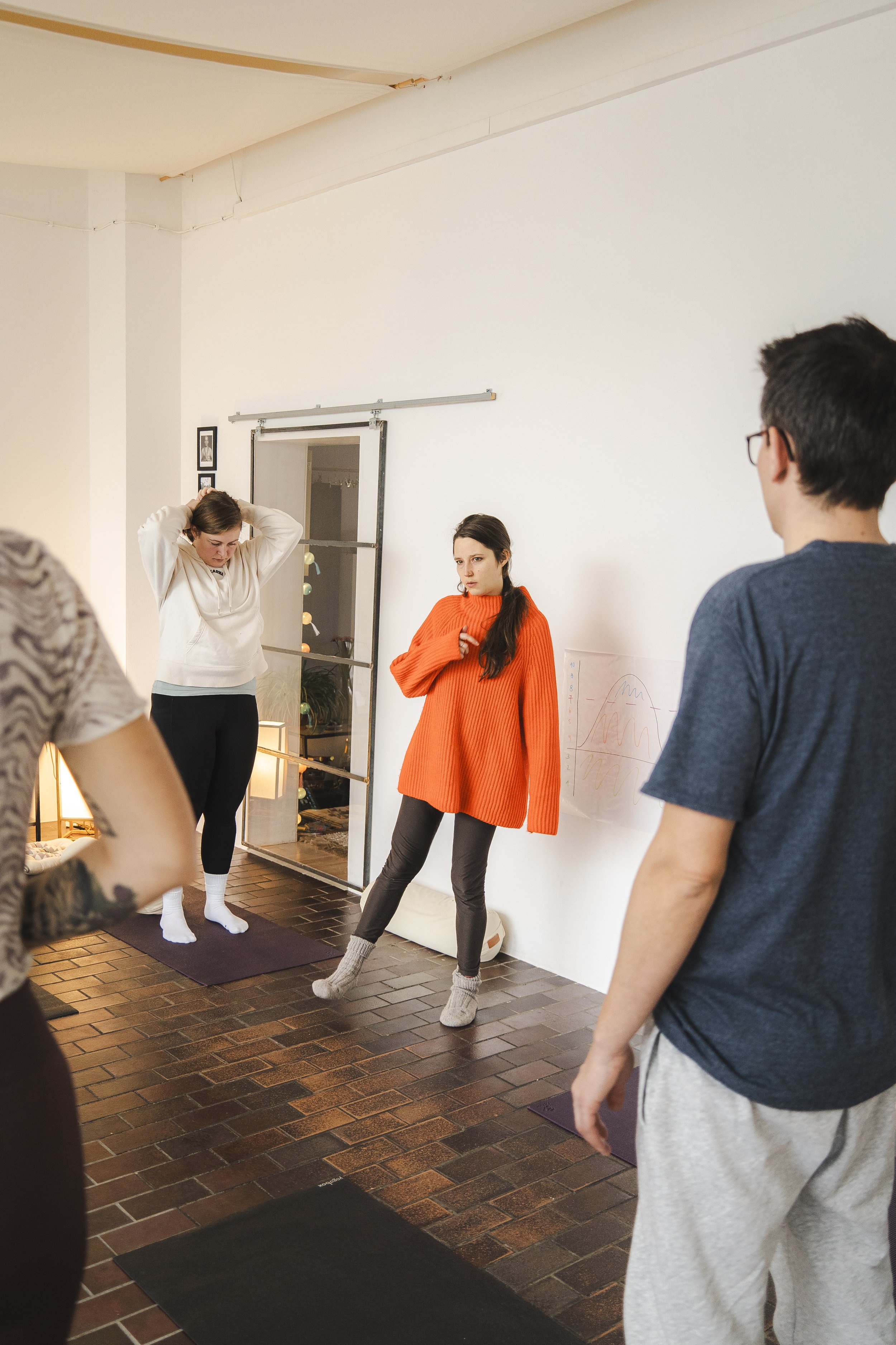 Gruppe von Menschen, die in einem hellen Raum stehen, möglicherweise an einem Yoga- oder Entspannungskurs teilnehmen. Eine Person in einem orangefarbenen Pullover spricht zu den anderen.