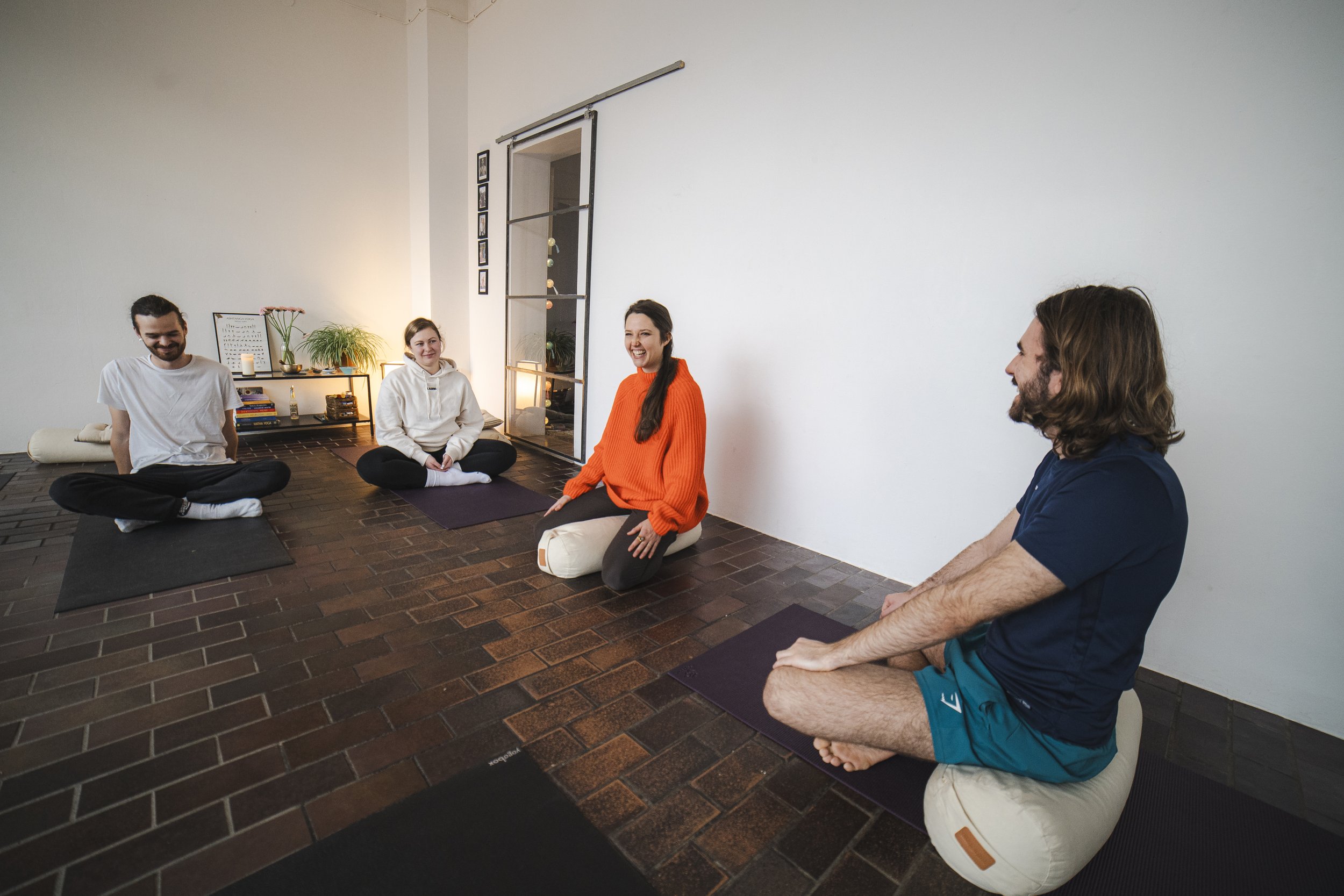Gruppe von vier Personen sitzt auf Yoga-Matten in einem Raum mit hellem Hintergrund, Pflanzen und Dekorationselementen, scheinbar in einer Meditations- oder Yogastunde.