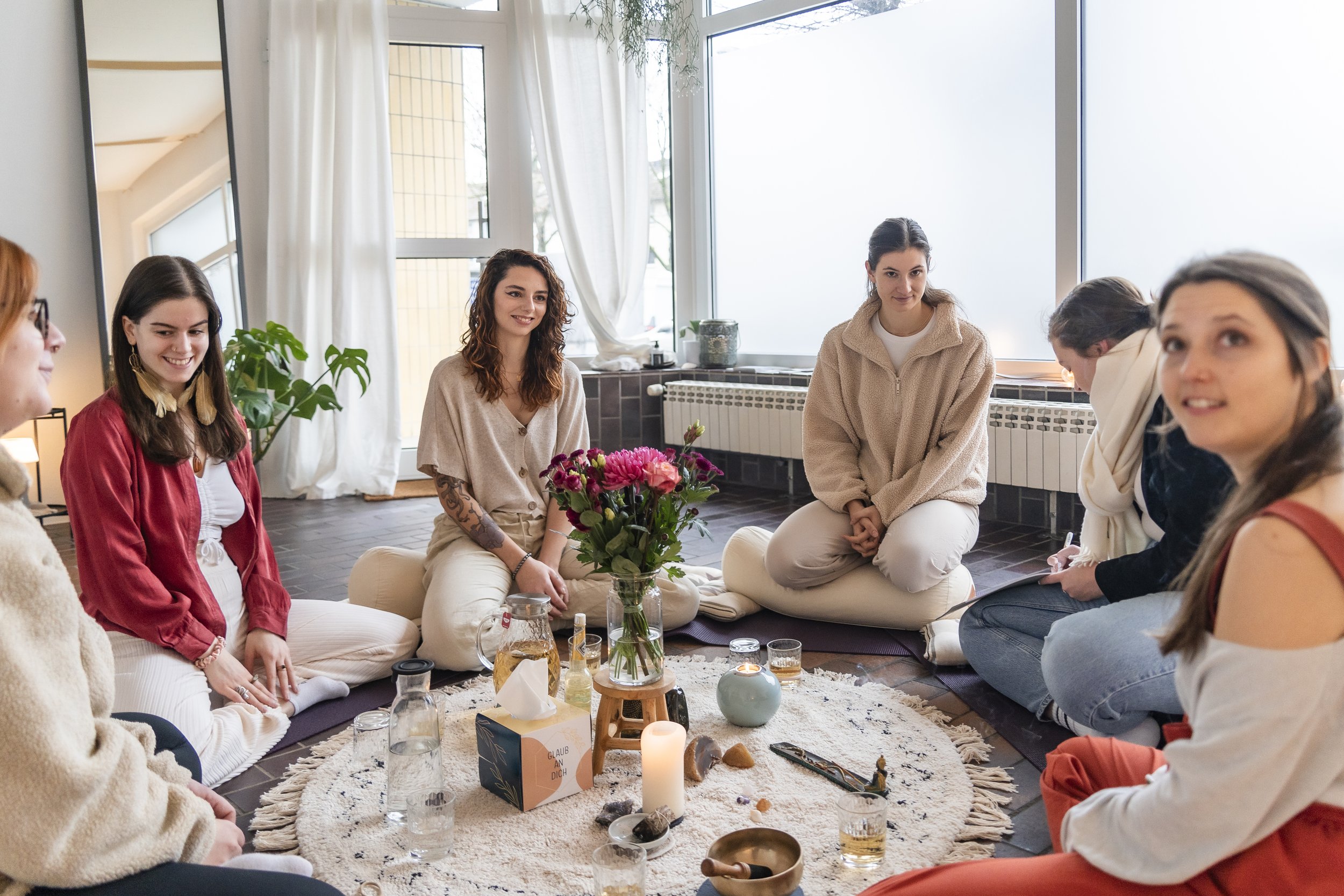 Eine Gruppe von sechs Frauen sitzt im Frauenzirkel. In der Mitte des Kreises befindet sich ein runder Teppich mit Dekorationen wie Blumen, Kerzen und Getränken.