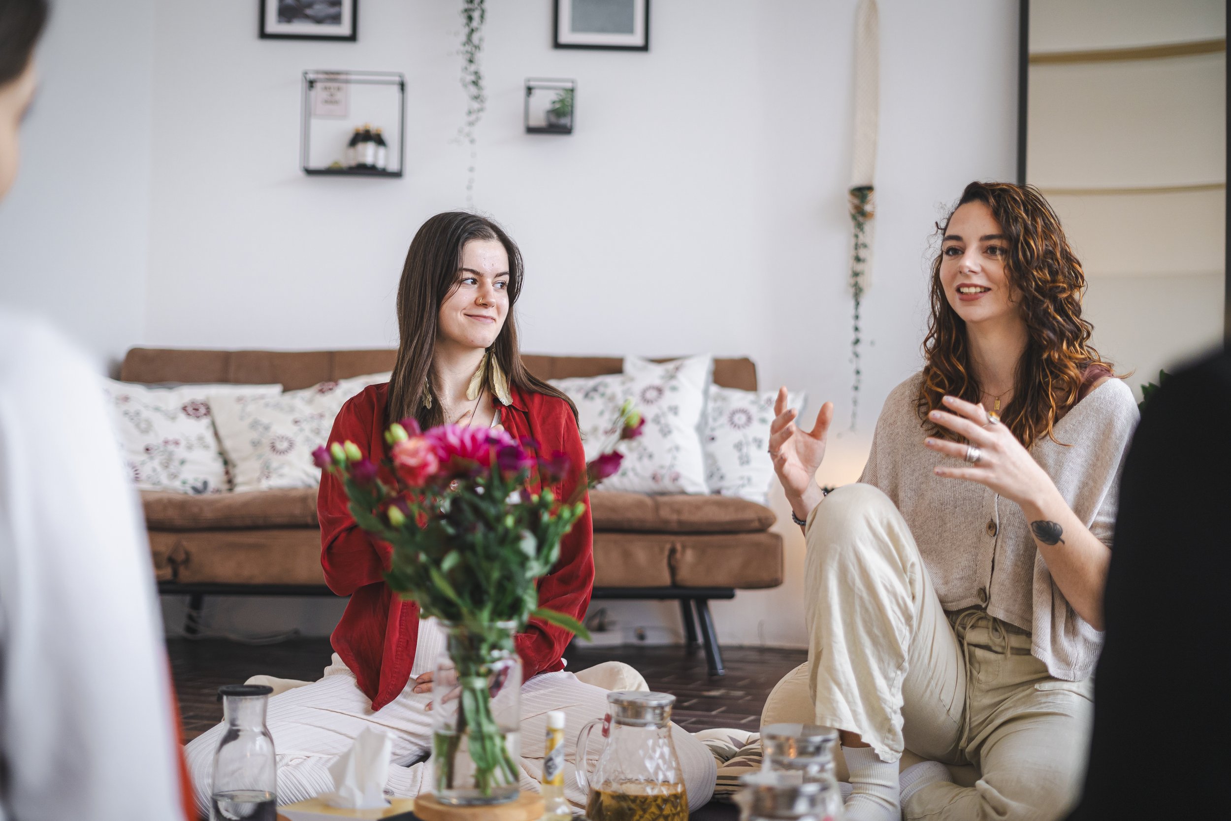 Zwei junge Frauen sitzen auf dem Boden und unterhalten sich. Im Vordergrund steht eine Vase mit Blumen. Im Hintergrund sind ein Sofa und eingerahmte Bilder an der Wand zu sehen.