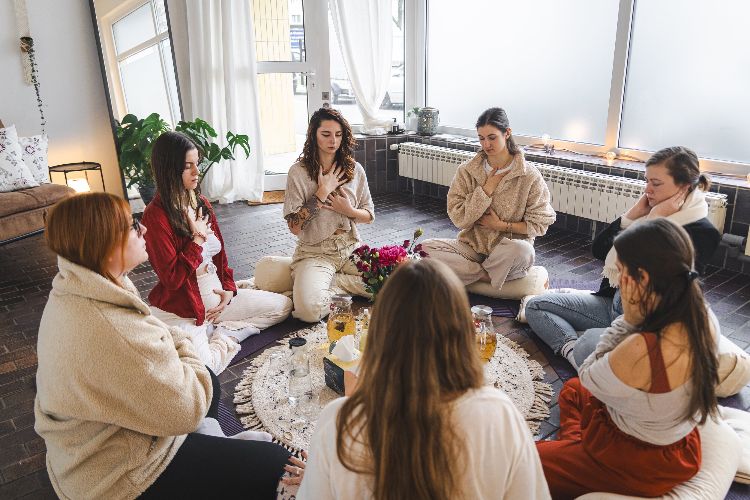 Gruppe von Frauen sitzt im Kreis, meditiert, in einem gemütlichen Raum mit Teppich und Blumen in der Mitte.