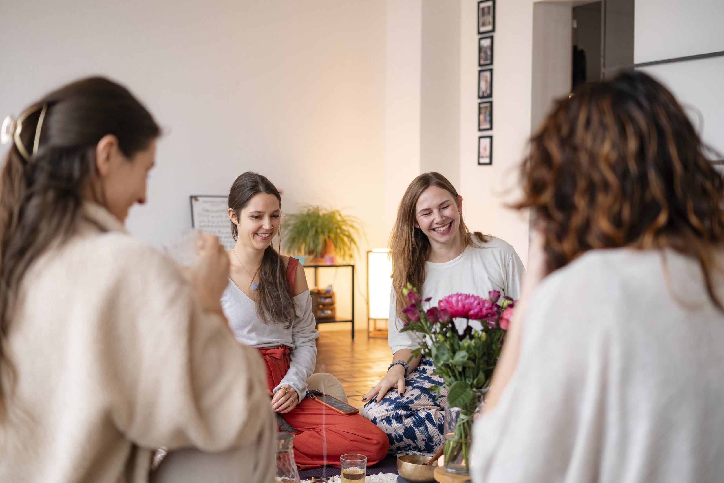 Vier Frauen sitzen auf dem Boden in einem Frauenzirkel und unterhalten sich fröhlich. Vor ihnen steht ein Tisch mit Blumen und Getränken.