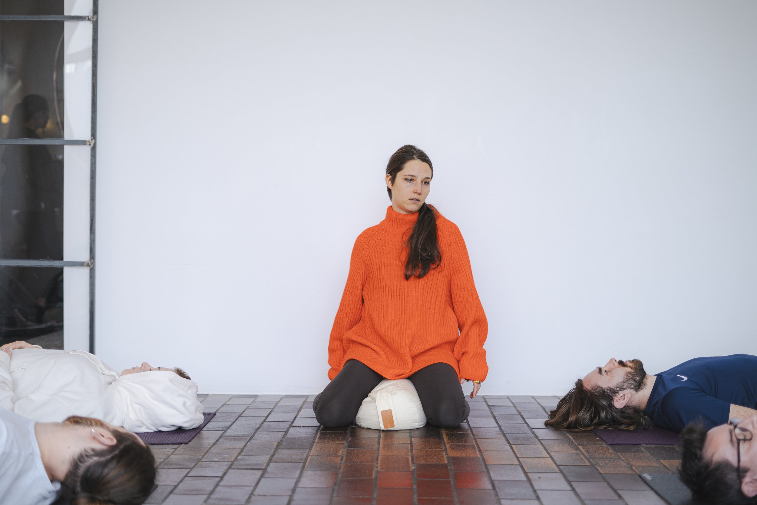 Eine Person in einem orangefarbenen Pullover sitzt meditierend auf einem Kissen, umgeben von mehreren Personen, die auf dem Boden auf Yogamatten liegen.